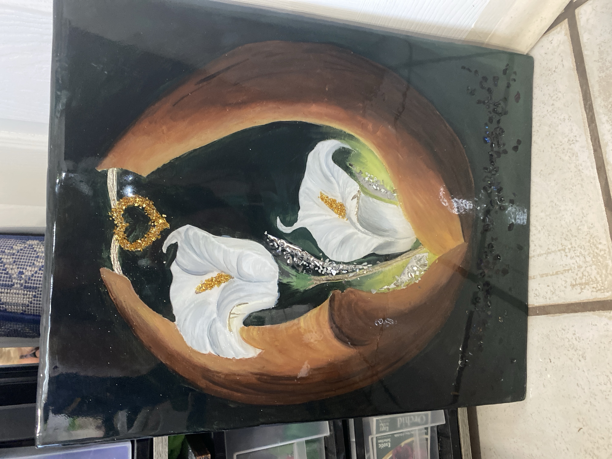 The image shows a glossy, rectangular artwork featuring two white calla lilies with golden centers, framed by two curving brown shapes. At the top, a small, gold, heart-shaped embellishment with glitter is suspended between the brown shapes. The background is dark, with subtle sparkles. The bottom edge of the artwork has some dark, irregular flecks and a few scattered iridescent sparkles. The artwork appears to be mounted on a black surface, possibly a frame or the backing of the piece itself. The artwork is propped up against a white baseboard, and a tiled floor is visible in the foreground. To the left of the artwork, a black shelving unit with some plastic cases containing what appear to be collectible cards is partially visible. The image is taken indoors. The city of Miami is not depicted in the image.
