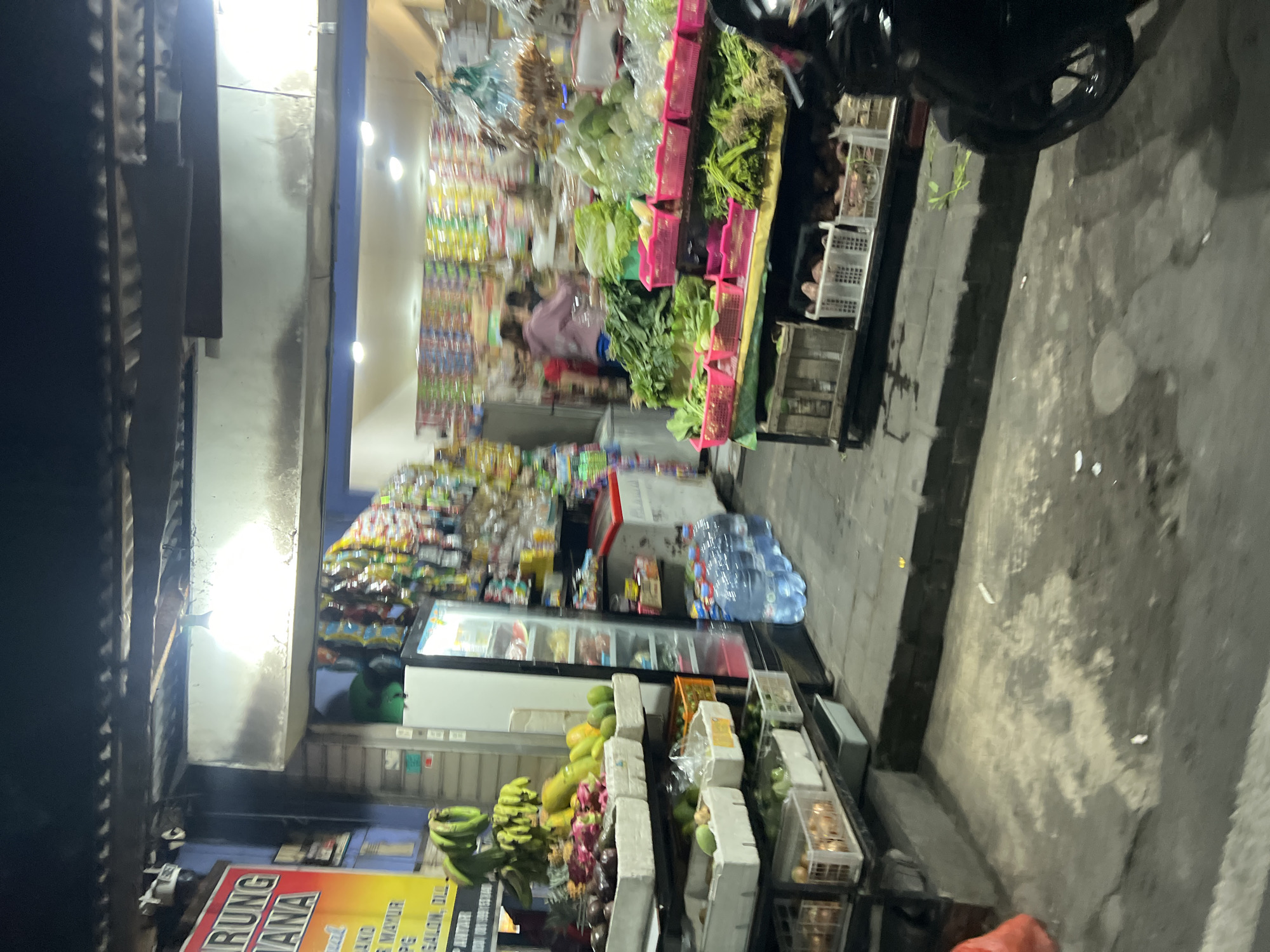 The image depicts a street-level view of a small market stall in Kuta, Indonesia, at night. The stall primarily sells fruits and vegetables. On the left, a sign with "ARUNG JANA" and other text is visible. Several bunches of green bananas hang prominently. Below, various fruits and vegetables are displayed in white crates and baskets, including what appear to be avocados, rambutans, papayas, and pineapples. A refrigerator unit is positioned behind the fruit display, with bottles of water lined up in front of it. To the right, more produce is arranged on tables and shelves, with a variety of leafy greens, cabbages, and root vegetables in pink baskets. Further back, shelves are stocked with packaged goods. Two individuals are partially visible inside the stall, one in a pink top. A motorcycle is parked to the right foreground. The ground is paved and appears wet. The overall lighting suggests it is nighttime, with artificial lights illuminating the stall.