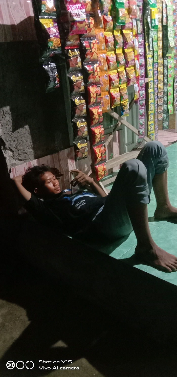 In Jabung Sisir, Indonesia, a single male individual is depicted resting on a light green tiled floor within what appears to be a small retail space. He lies on his back, wearing a dark t-shirt with a graphic print and grey trousers, with his feet bare. His left hand is positioned behind his head, while his right hand holds a white smartphone, towards which his gaze is directed. To the right of the individual, a vertical display system is visible, densely packed with numerous colorful packaged snack items, including brands like "Chitato." A rough-textured wall or column is present on the far left. Strong, directional artificial light illuminates the scene, creating defined shadows on the floor and the left wall.