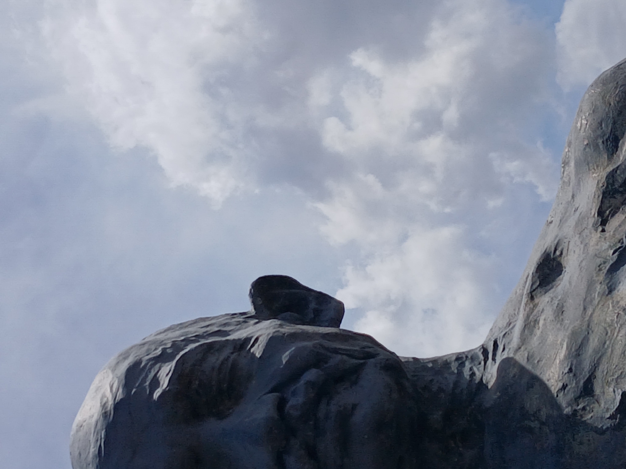 The image shows a statue against a cloudy sky. The statue appears to be made of dark-colored stone or metal and has a rough, textured surface. The subject matter is obscured, but it might be a mountain or a figure with a prominent head or headdress. Given the mention of Ciudad Guayana, Venezuela, the statue is likely the Monumento a la CVG, a well-known landmark.