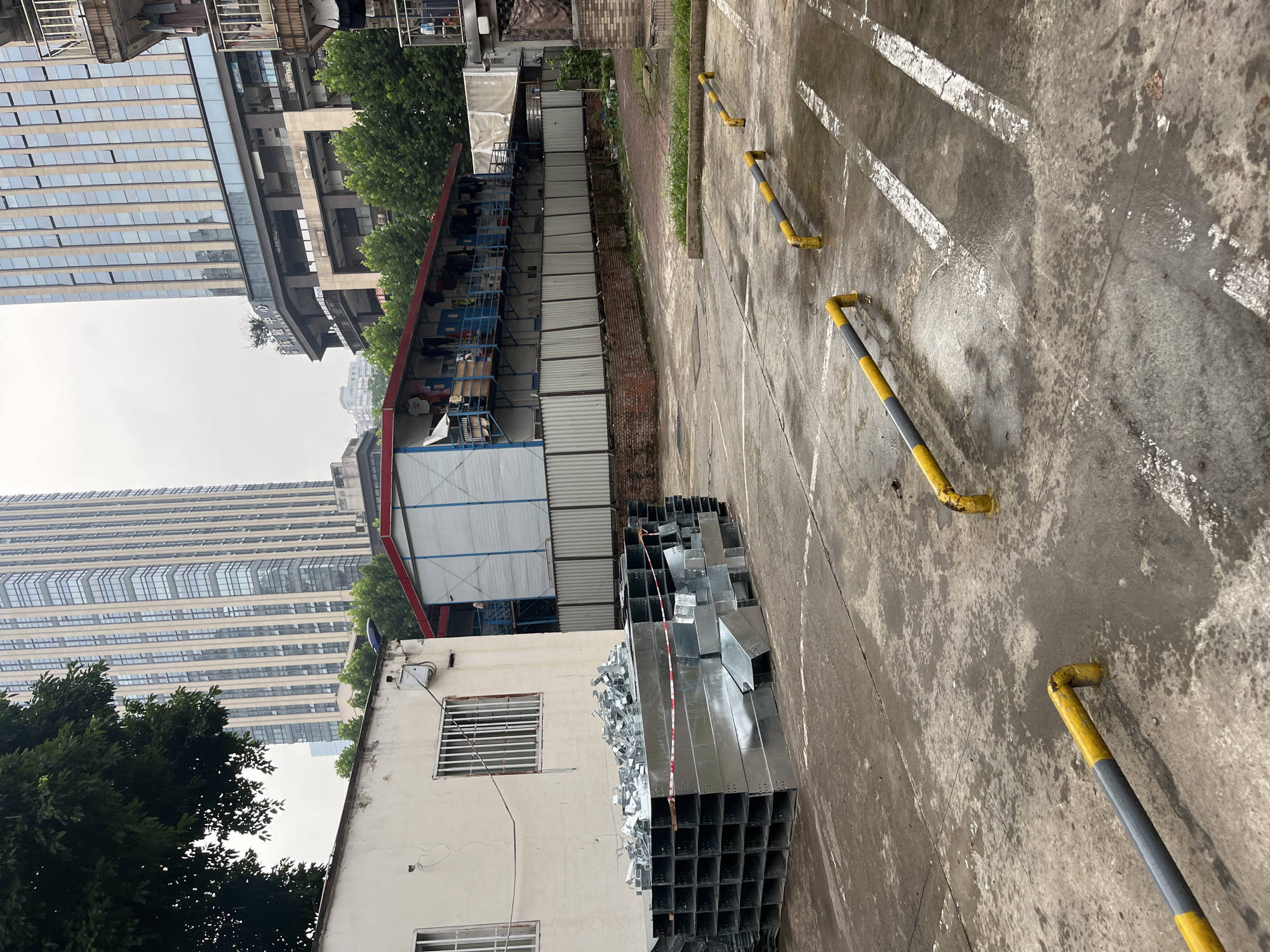 In the Yuzhong District of China, this outdoor scene depicts a temporary construction site or storage area. In the foreground, a wet, concrete surface shows signs of wear and tear, with painted white lines that appear to delineate parking spots. Several yellow and gray metal bollards are scattered across the pavement, some bent or damaged. A large pile of stacked galvanized metal ducts or channels is positioned on the left side of the frame. Behind this, a low, white, single-story building with barred windows stands. Further back, a modular temporary structure with a blue railing and what appear to be laundry hanging from it is visible. Beyond this, several tall, modern buildings dominate the skyline, with a large, glassy skyscraper in the background. Green trees are interspersed between the structures. The sky is overcast, suggesting a cloudy or possibly rainy day.