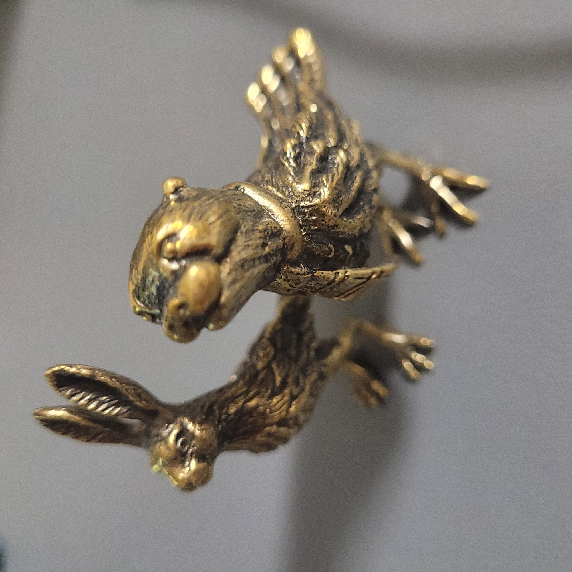 This is a close-up, high-angle, shallow depth of field photograph of two small, metallic animal figurines, appearing to be made of brass or a similar alloy. The figurine on the left is a rabbit, facing left, with its long ears held upright. Its body is elongated, and its hind legs are extended backward. The figurine on the right is a bird, facing right and slightly upward. It has outstretched wings, small feet, and its head is tilted as if it is looking at something. The figurines are placed on a smooth, solid gray surface. The background is also a soft, out-of-focus gray. The lighting casts a warm glow on the metallic surfaces, highlighting the textured details of the animals' fur and feathers. The overall impression is one of a still life composition, emphasizing the craftsmanship of the figurines.