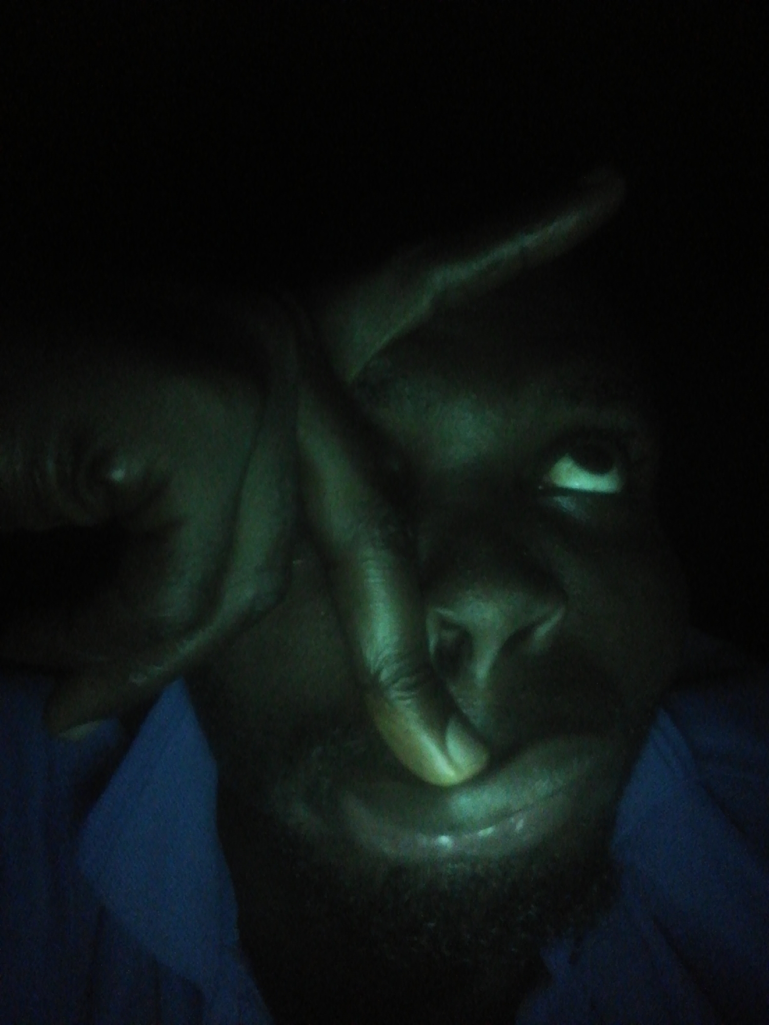 A dimly lit close-up photograph depicts a dark-skinned individual with a beard and blue shirt, partially obscuring their face with their hand, forming a shape over their eye. The location is unknown.