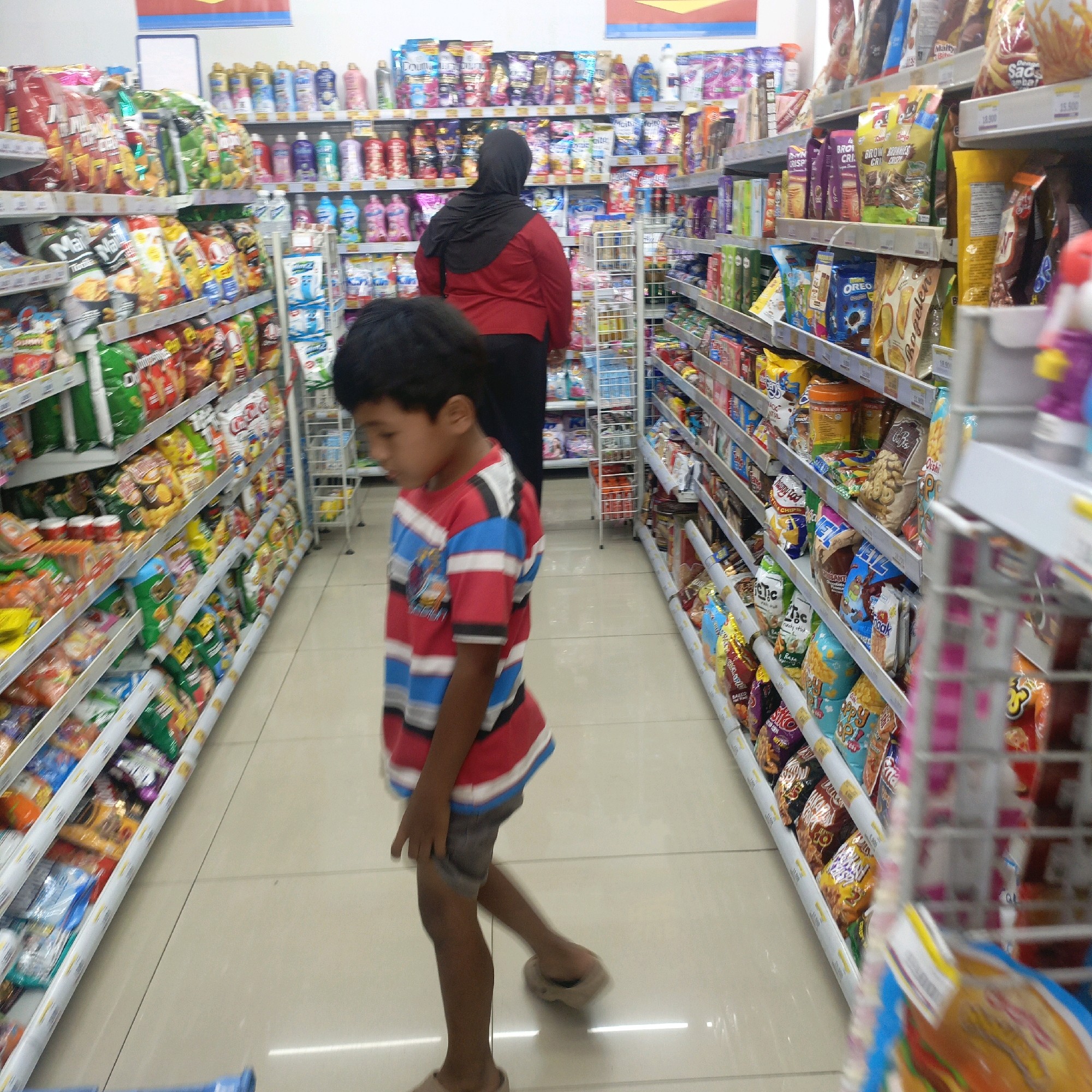 This is a medium-shot, slightly high-angle, indoor photograph of a convenience store aisle in Salatiga, Indonesia. The aisle is filled with shelves of packaged goods, primarily snacks and toiletries. In the foreground, a young boy wearing a red, blue, and white striped shirt and grey shorts is walking away from the camera and towards the right side of the frame. His right leg is extended, and his left foot is planted on the floor. Behind the boy and to the left, a woman wearing a black hijab and a red shirt is standing, facing away from the camera. The shelves are stocked with a variety of colorful snack bags, bottles, and boxes. The floor is tiled in a light beige color.
