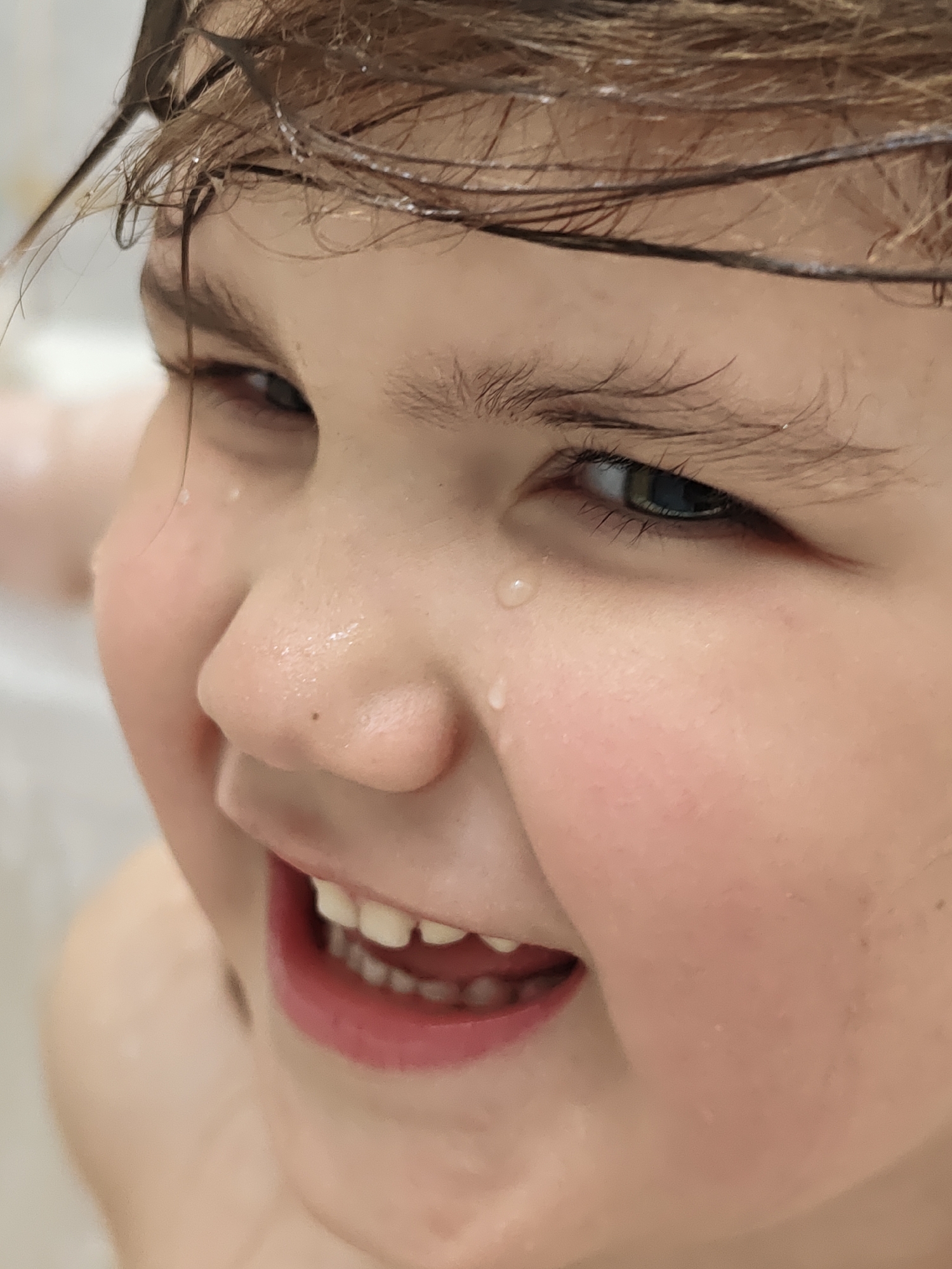 The image captures a close-up, high-angle shot of a young child, likely of pre-school age, with wet, light brown hair. The child's face is the primary focus, radiating joy and amusement. Their eyes, a striking blue or green, sparkle with delight, and their gaze is directed upwards and to the right, suggesting they are looking at something engaging. A wide, genuine smile reveals several upper and lower teeth, some of which appear to be baby teeth with small gaps, indicating a young age.
Numerous water droplets cling to the child's wet hair and skin, particularly around the nose, cheeks, and below the left eye, where a prominent tear-shaped drop rests. These droplets suggest the child is actively playing in or has just emerged from water, possibly a bath or shower. The background is softly blurred, showing indistinct light tones that could be a bathroom environment, like a tub or tiled wall, further supporting the idea of a water-related activity.
The dominant emotion conveyed is one of pure happiness and playfulness. The image captures a fleeting moment of childlike exuberance. There is no visible text. Given the location context of Chaikovsky, Russia, the scene depicts a universal moment of childhood joy, potentially during a bath or water play at home.