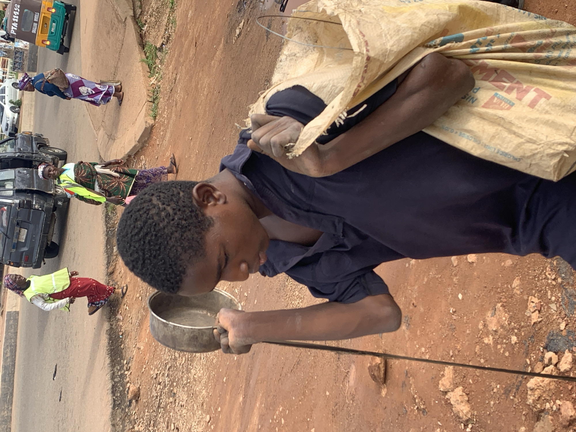 A young male, appearing to be a teenager, walks on a dirt path beside a road. He is carrying a large, light-colored sack over his right shoulder. The sack appears to be made of rough material and has some red lettering visible, possibly indicating cement. In his left hand, he holds a metal pot or bowl by a long handle. He is wearing a dark blue collared shirt. In the background, on the paved road, three women are visible. One woman in the foreground wears a bright yellow vest over a red and white patterned outfit and walks away from the camera. Further down the road, two other women are standing on a raised concrete area. One is wearing a purple and white patterned dress and headscarf, and the other is wearing a green and white patterned wrap with a yellow vest, carrying a baby on her back. Various vehicles are present on the road, including a black van, a white car, and a green and yellow auto-rickshaw. The environment appears to be an urban or semi-urban area in Gura Top, Nigeria, characterized by a dirt road, a paved road, and buildings in the distance. The ground is a reddish-brown dirt with scattered rocks and debris.