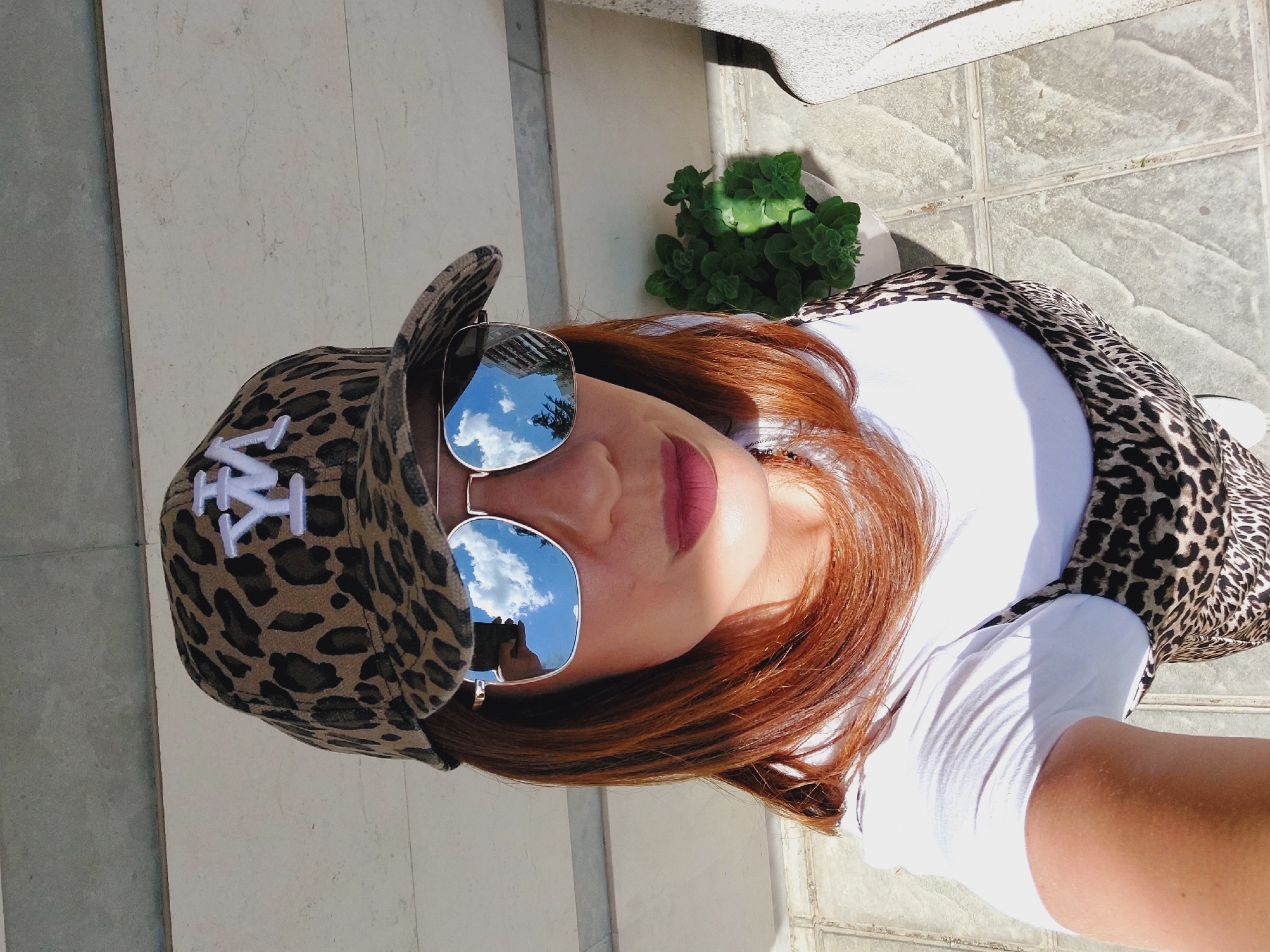 A woman with reddish-brown hair is shown from a high angle, wearing a leopard-print baseball cap with a white "YW" logo, aviator sunglasses reflecting a bright blue sky with clouds and buildings, a white t-shirt, and a leopard-print slip dress. She is standing on a tiled outdoor surface, possibly a patio or walkway. To her right, a potted green plant is visible. The background shows a concrete structure, consistent with an urban environment. The image was taken in Sofia, Bulgaria.