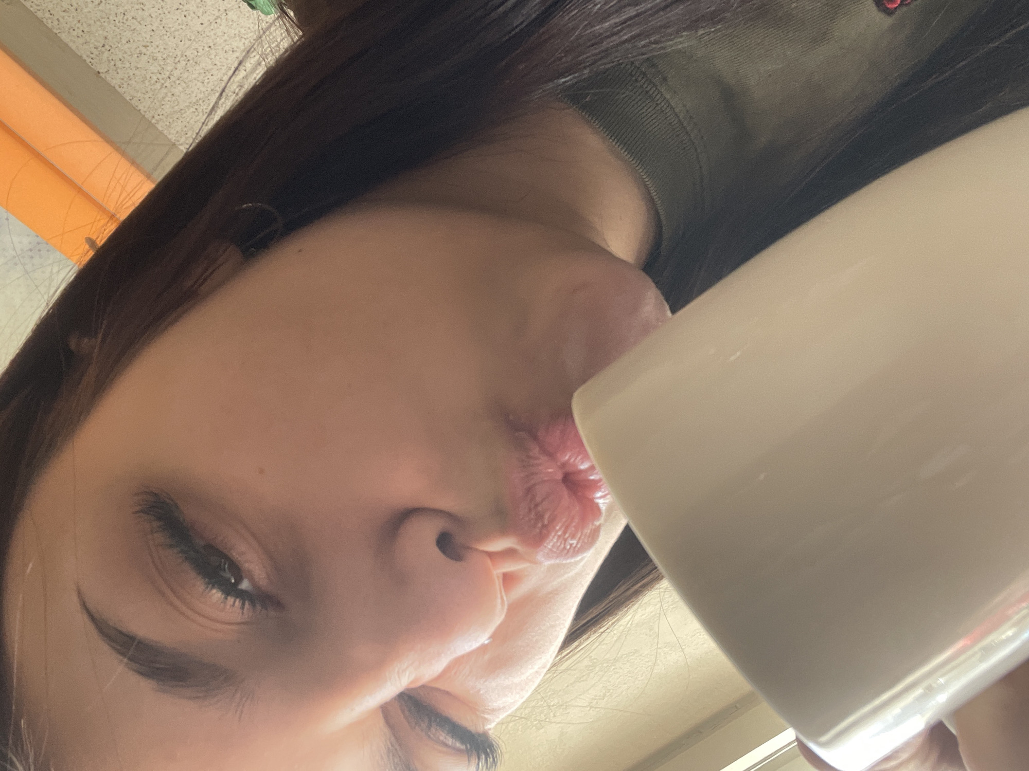 A close-up, angled selfie captures a young woman with dark brown hair. Her lips are pursed into a kiss. She is holding a white mug. The background reveals glimpses of orange tubes and a speckled surface, suggesting an interior space. The location appears to be in a city but there is no information available to specify Saint Petersburg, Russia.
