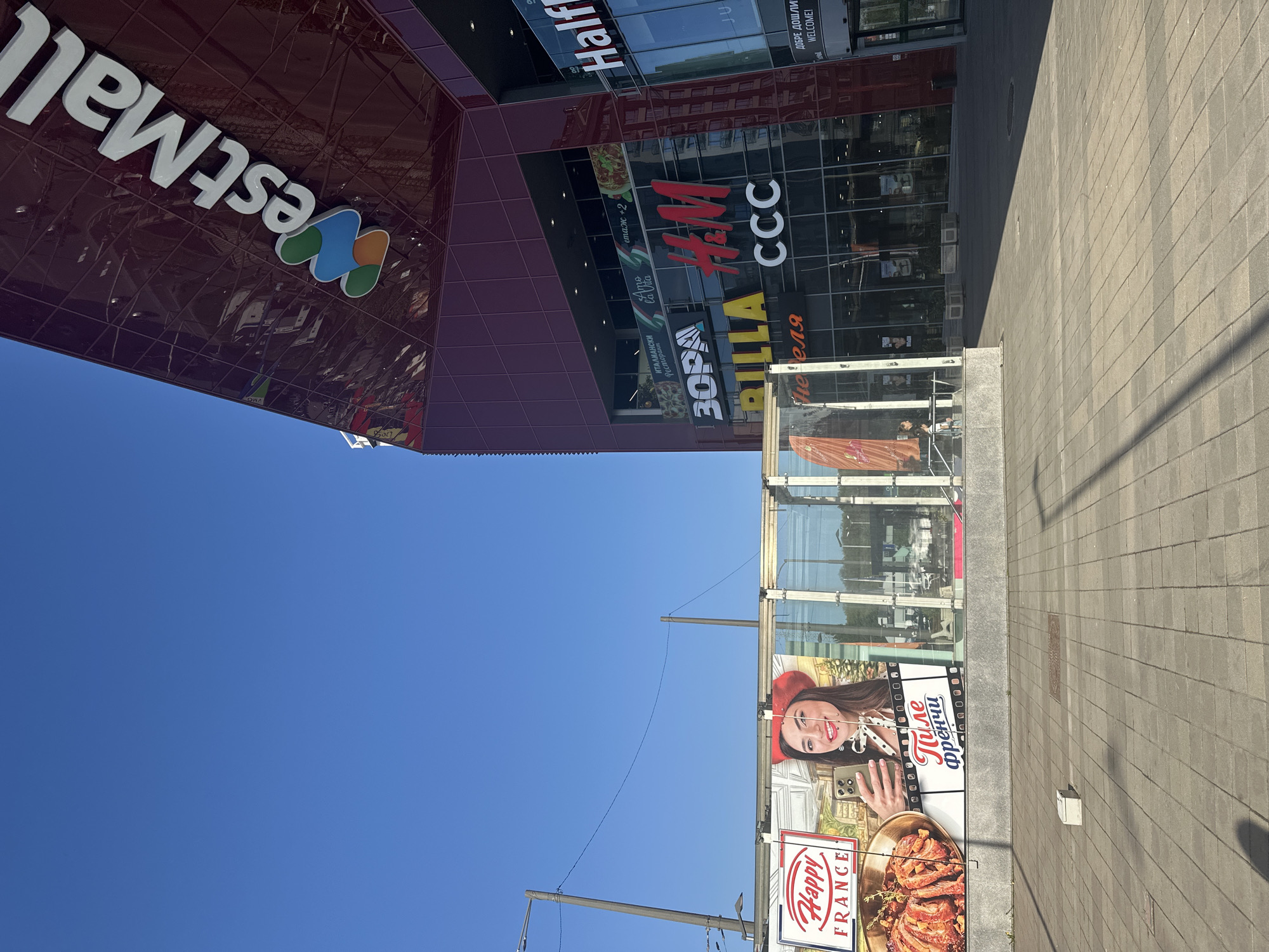 This image captures the exterior of a modern shopping mall and the surrounding urban environment under a clear blue sky. The main subject is the "West Mall" building, with its name prominently displayed in white letters and a colorful, abstract logo. The mall's facade features large glass windows reflecting the surroundings, and several store logos are visible, including "Half," "H&M," "CCC," and "BILLA." Other Cyrillic text identifies businesses like "ЗОРА" (Zora), "ИТАЛИАНСКИ Ресторант" (Italian Restaurant), and "етаж +2" (floor +2). A welcome sign in English and Bulgarian reads "ДОБРЕ ДОШЛИ! WELCOME!".
The setting is an outdoor area, likely a pedestrian walkway or entrance to the mall. The ground is paved with light-colored square tiles, and shadows suggest it's daytime with bright sunlight. To the right, a large billboard features a smiling woman holding a smartphone, with the text "Пиле Френчи" (Chicken Frenchy) and "Happy FRANCE" next to an image of a cooked dish. The sky is a vibrant, cloudless blue, occupying a significant portion of the upper part of the image. The overall impression is a bustling commercial area on a bright, sunny day in Sofia, Bulgaria, as indicated by the Cyrillic script. No specific activities are ongoing, but the scene implies a commercial and potentially active area.
