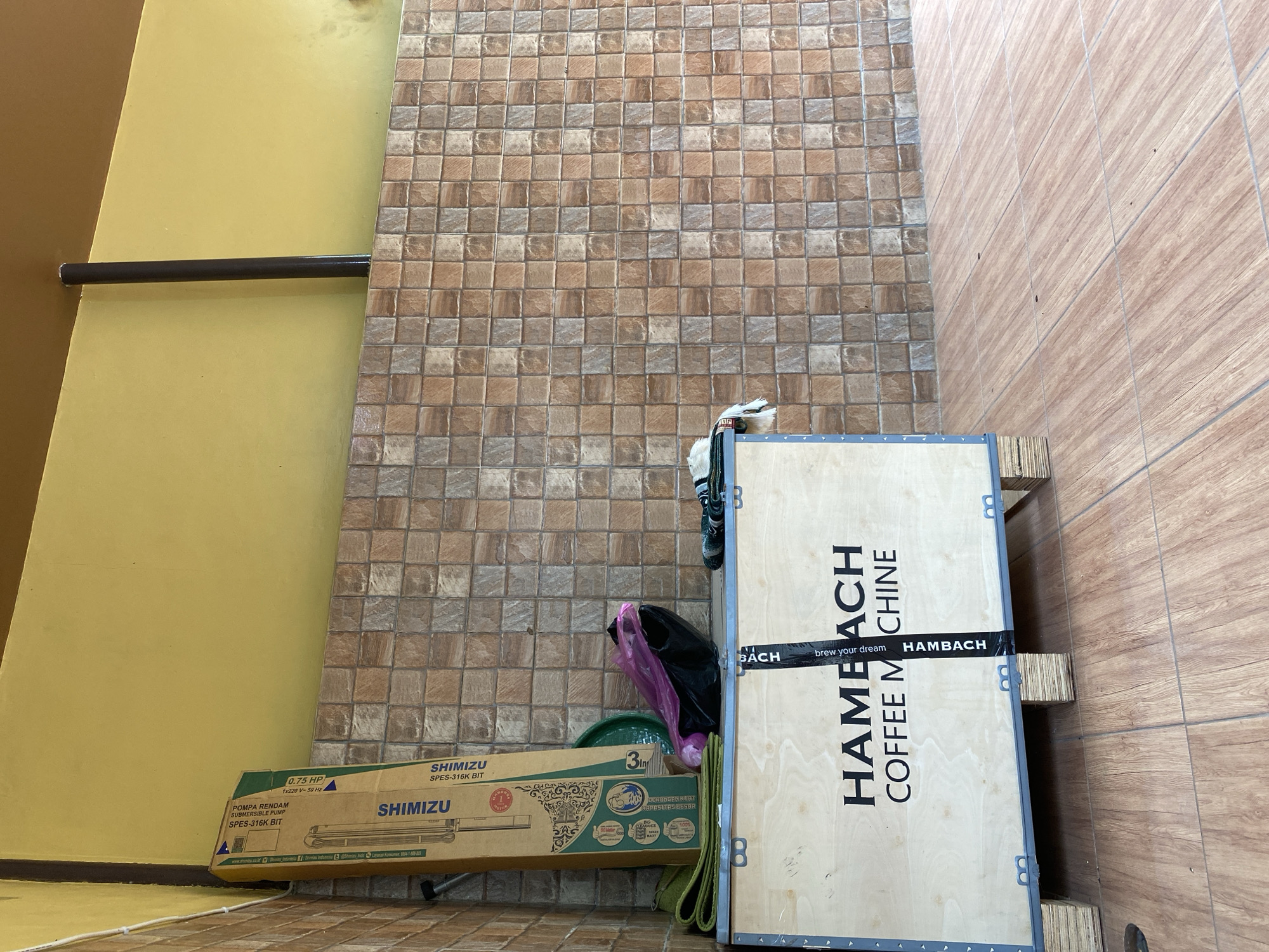 The image depicts an outdoor, covered area with tiled flooring and a tiled wall. In the foreground, a large wooden crate is prominently displayed, bearing the branding "HAMBACH COFFEE MACHINE." To the left of the crate, a tall cardboard box, identified as a "SHIMIZU SPES-316K BIT SUBMERSIBLE PUMP," is leaning against the wall. Next to the pump box, a green plastic basin and a purple plastic bag containing what appear to be fabric items are visible. A dark brown pipe extends vertically from the ceiling down towards the wall in the middle ground. The overall scene suggests a storage or staging area, possibly in a commercial or industrial setting in Tangerang, Indonesia, given the provided context.