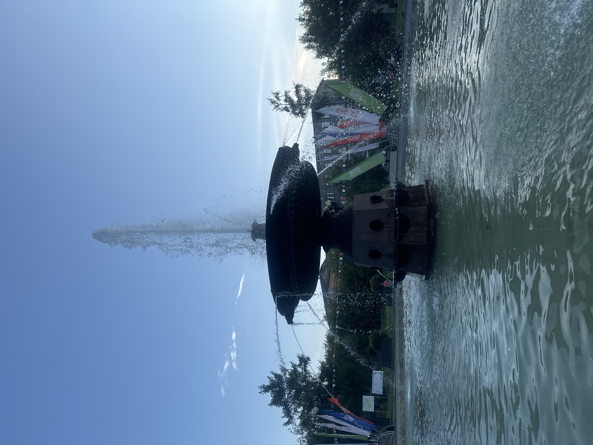 The image captures a large, ornate fountain in a city square, likely in Irkutsk, Russia, given the presence of national flags. The fountain is made of dark stone and features multiple tiers, with water cascading from the upper basin and a powerful jet shooting upwards. The water in the basin is a murky turquoise, reflecting the sky and surrounding greenery. Several flags, some appearing to be the Russian tricolor and others green with white symbols, are prominently displayed on poles around the fountain. In the background, dense green trees line the perimeter of the square, with a few buildings visible behind them, their architecture suggesting a historical or civic area. The sky is a clear, bright blue with a few wispy clouds, indicating daytime. The overall scene suggests a public space, possibly a park or a central plaza, that is active and maintained, as evidenced by the flowing water and the festive decorations.