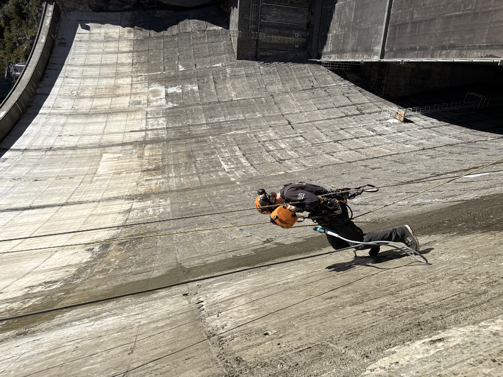 A construction worker is rappelling down the face of the dam in Castellane, France. He is wearing a helmet and safety gear. He is suspended by ropes and is using a tool to work on the dam face. The dam is made of concrete.