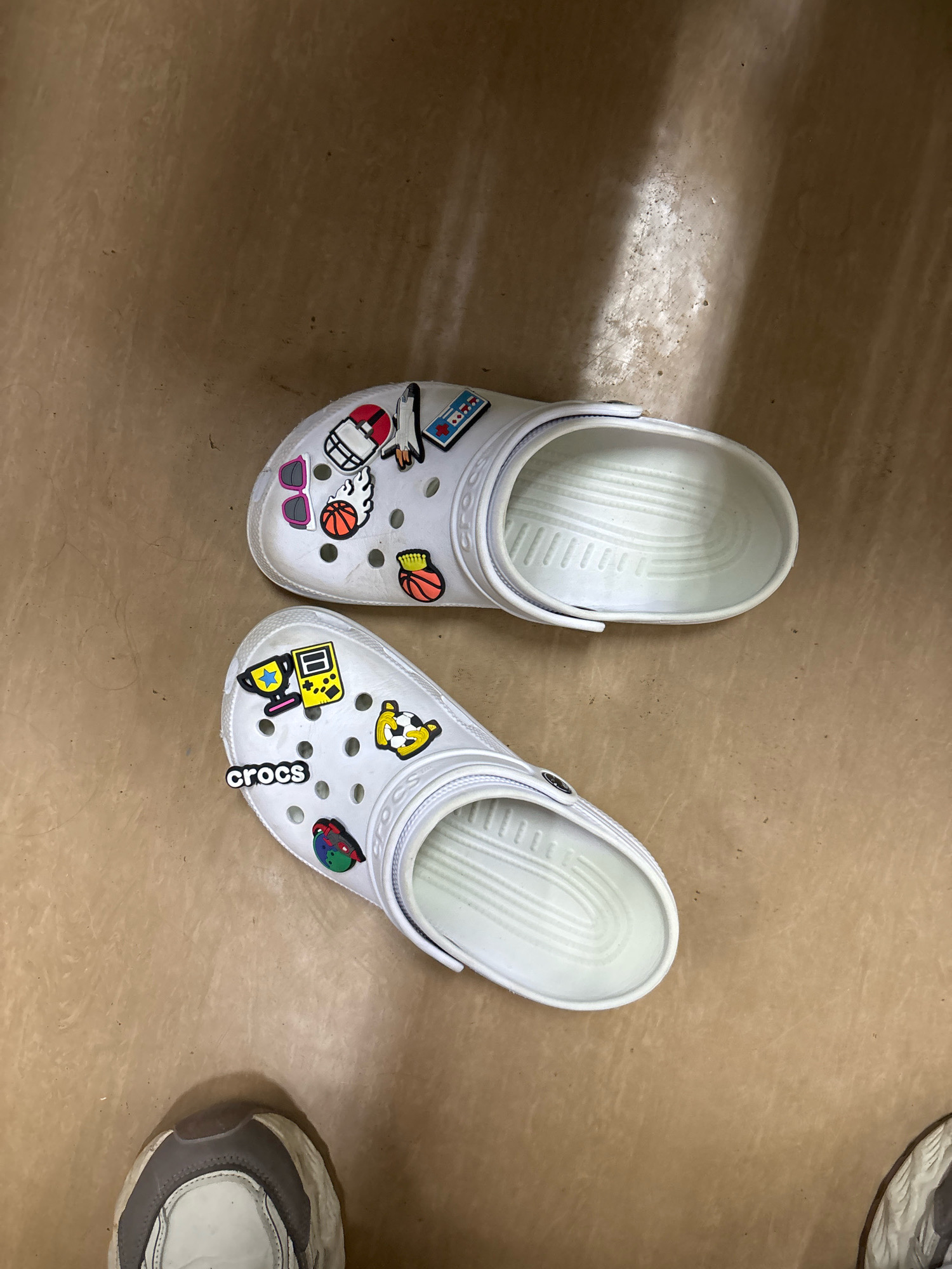 A pair of white Crocs adorned with various colorful charms sits on a brown, textured surface. The charms on the top Croc include a rocket, a basketball, sunglasses, a video game controller, and a football helmet. The bottom Croc features the word "crocs" in black, along with a trophy, a soccer ball, and a red, green, and blue circular charm. The surface appears to be flooring or a similar material. The lighting creates shadows on the surface, suggesting an indoor environment. A portion of a grey and white sneaker is visible in the lower left corner of the image, and another sneaker is partially visible in the lower right. The image is taken from a top-down perspective. There is no indication of the location in Chaikovsky, Russia, or any people interacting with the Crocs.