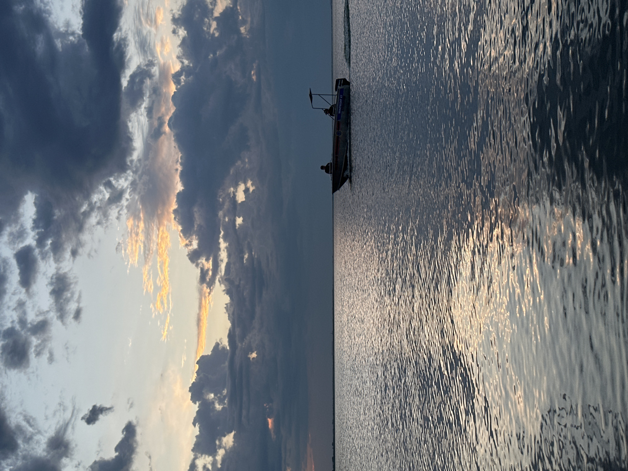 The image depicts a serene water scene at dusk, with a boat moving across a calm body of water. The sky is filled with dramatic clouds, tinged with warm orange and yellow hues from the setting sun, contrasting with the darker, moody blues and grays of the storm clouds. The water reflects the sky's colors, creating shimmering patterns. In the mid-ground, a small boat with at least two silhouetted figures is visible. The boat appears to be a motorboat, with a wake trailing behind it, indicating movement. The horizon line is low, separating the vast expanse of water from the expansive sky. The overall atmosphere is one of tranquility and the dramatic beauty of nature. The location is identified as the city of Kokshetau, Kazakhstan.