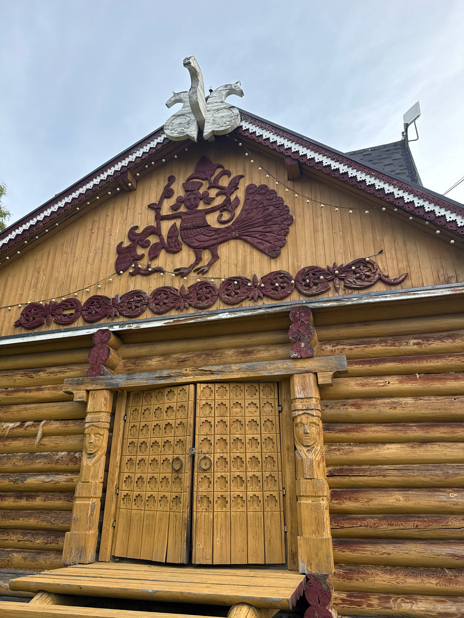 The image shows the facade of a wooden building, likely a traditional Russian izba, in Peterhof, Russia. The building is constructed from large, horizontal logs. The roofline is decorated with intricate wooden carvings, including a large bird-like figure on the apex and a band of floral motifs below. There is also a row of small, light bulbs strung along the upper part of the facade.
The entrance features a pair of wooden doors with a geometric pattern carved into them. The door frame is adorned with carved figures on either side. A wooden porch with a simple railing leads up to the doors. The overall impression is of a rustic, folk-art inspired structure. The sky above is overcast. There are no people visible in the image.