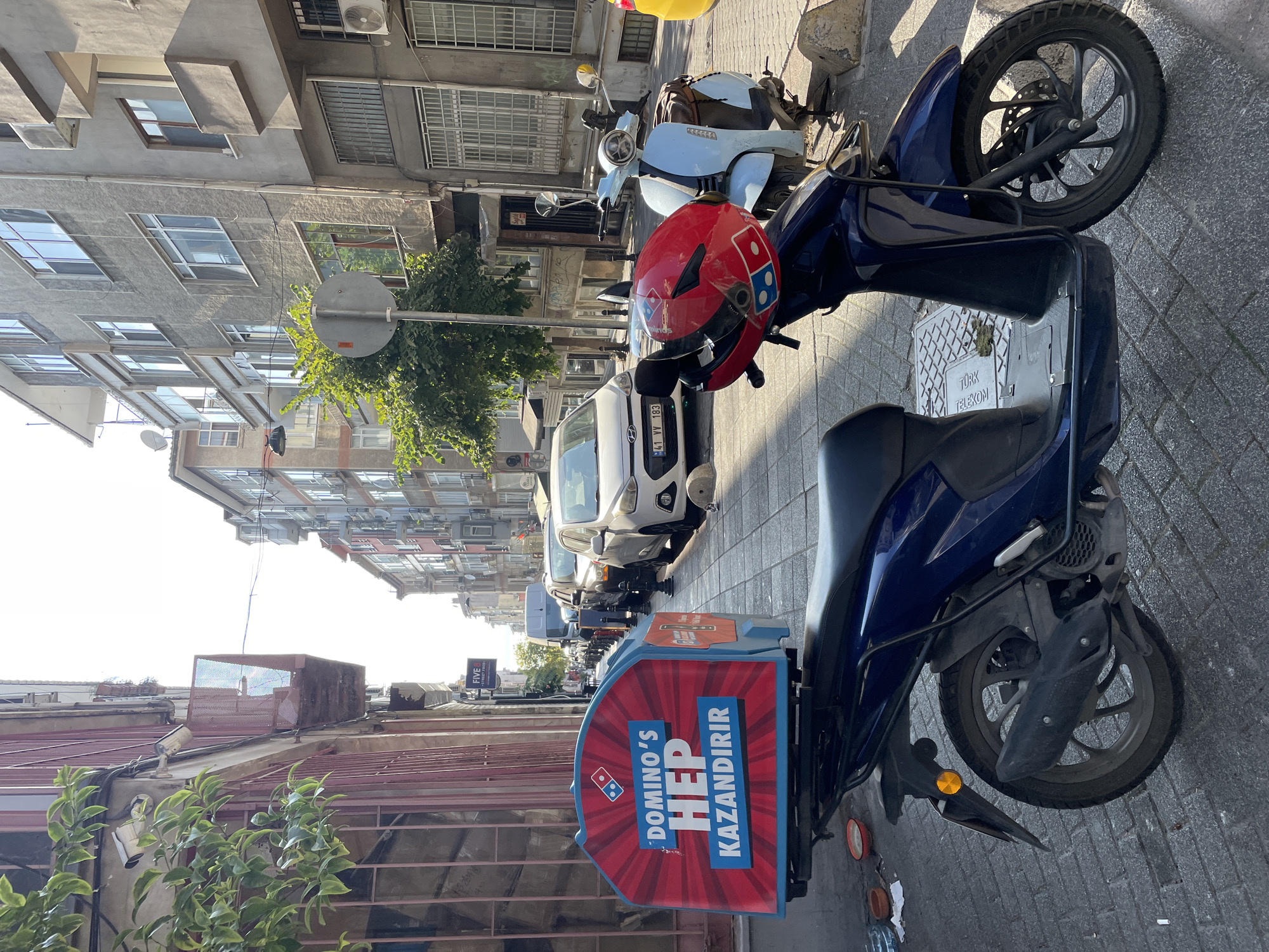 A dark blue scooter with a Domino's Pizza delivery box is parked on a cobblestone street in Kadıköy, Turkey. The delivery box is red with a white and blue Domino's logo and text that reads "DOMINO'S HEP KAZANDIRIR" in white and blue lettering. A red Domino's helmet rests on the handlebars of the scooter. In the background, there are several cars parked along the street and apartment buildings. A light blue scooter is parked next to the Domino's scooter. The street is lined with trees and buildings, suggesting an urban environment. The lighting indicates it is daytime, with sunlight casting shadows.