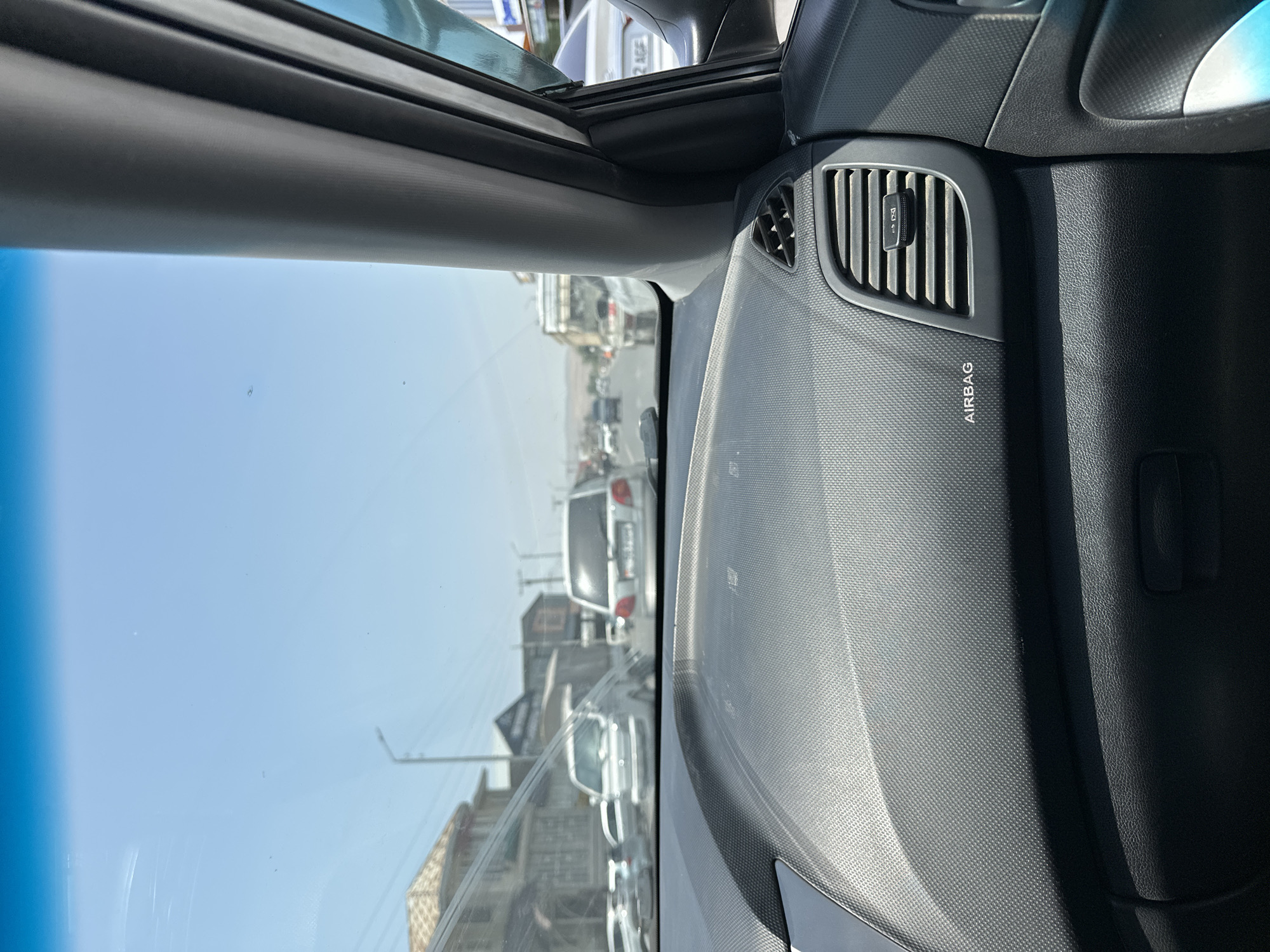 The image is taken from inside a car, looking out through the windshield at a city street. The dashboard is visible in the foreground, with an "AIRBAG" label and an air vent. Outside the car, there is traffic on a sunny day. Several cars are visible, including a white van in the middle distance and a white car in the foreground on the right. Buildings line the street, and there are power lines and street lights. The sky is blue with a few faint clouds. The scene appears to be a typical urban environment, likely in Manas, Kyrgyzstan, based on the context provided.