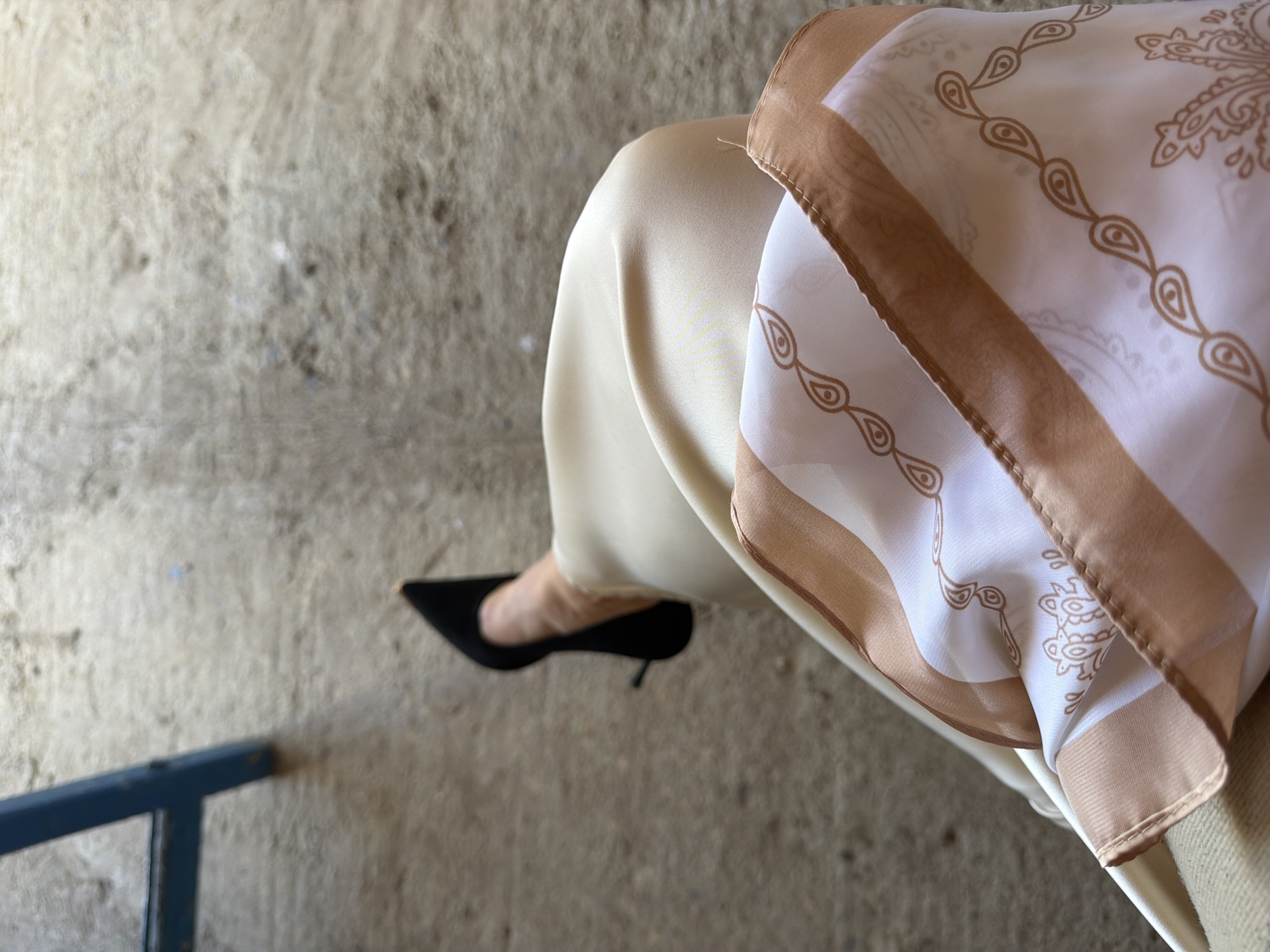 A low-angle, close-up shot shows a person's legs and feet, clad in black stiletto heels and a light-colored, flowing skirt with a patterned trim. The background is a rough, concrete surface, suggesting an outdoor or industrial setting. A portion of a blue metal chair leg is visible in the upper left corner. The overall lighting is natural, creating soft shadows. The scene is devoid of any context that would place it in Zăluceni, Moldova.