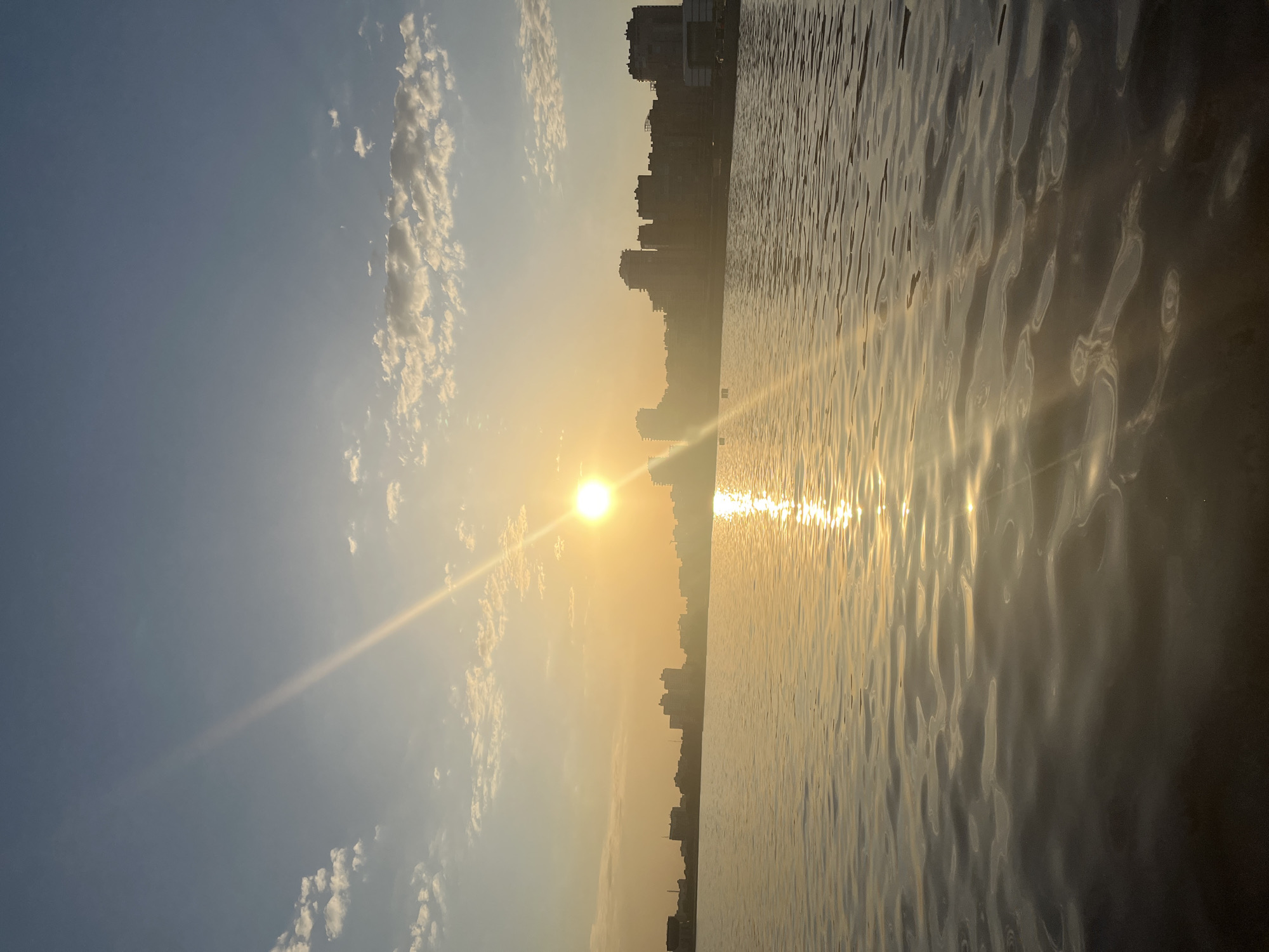 The image shows a sunset view over water, likely the Kazanka River in Kazan, Russia. A city skyline is silhouetted against the setting sun on the horizon. The sun reflects intensely on the water's surface, creating a bright, shimmering path. The sky transitions from a lighter blue with scattered clouds to a warmer orange near the horizon. The image appears to have been rotated, presenting the scene on its side.