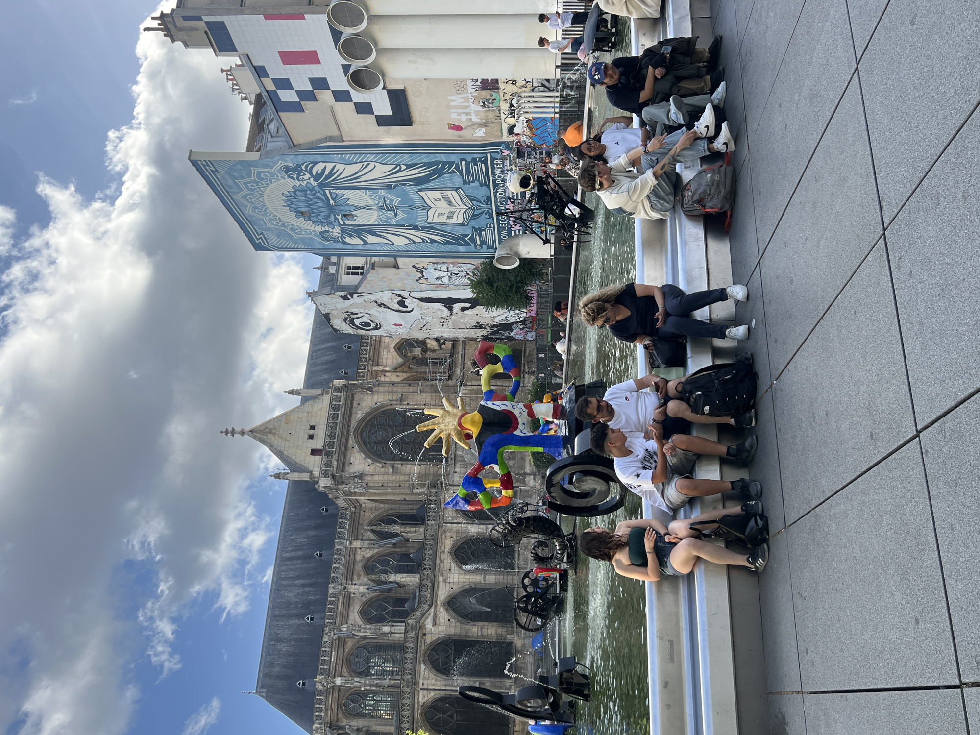 This image captures a vibrant urban scene in Paris, France, specifically at the Stravinsky Fountain. The fountain, a playful and colorful collection of abstract sculptures, is the central focus, with water jets spraying into a pool. Several people are seated on the edge of the fountain's basin, some engaged in conversation, others looking at their phones. They are a mix of adults and young people dressed in casual attire, suggesting a relaxed atmosphere.
Behind the fountain, the historic Saint-Merri Church stands majestically, its Gothic architecture contrasting with the modern art of the fountain and the surrounding buildings. These buildings are adorned with large, colorful murals and graffiti, adding to the artistic and dynamic feel of the location. The sky above is a mix of blue with scattered clouds. The overall scene depicts a lively public space where art, history, and everyday life converge.