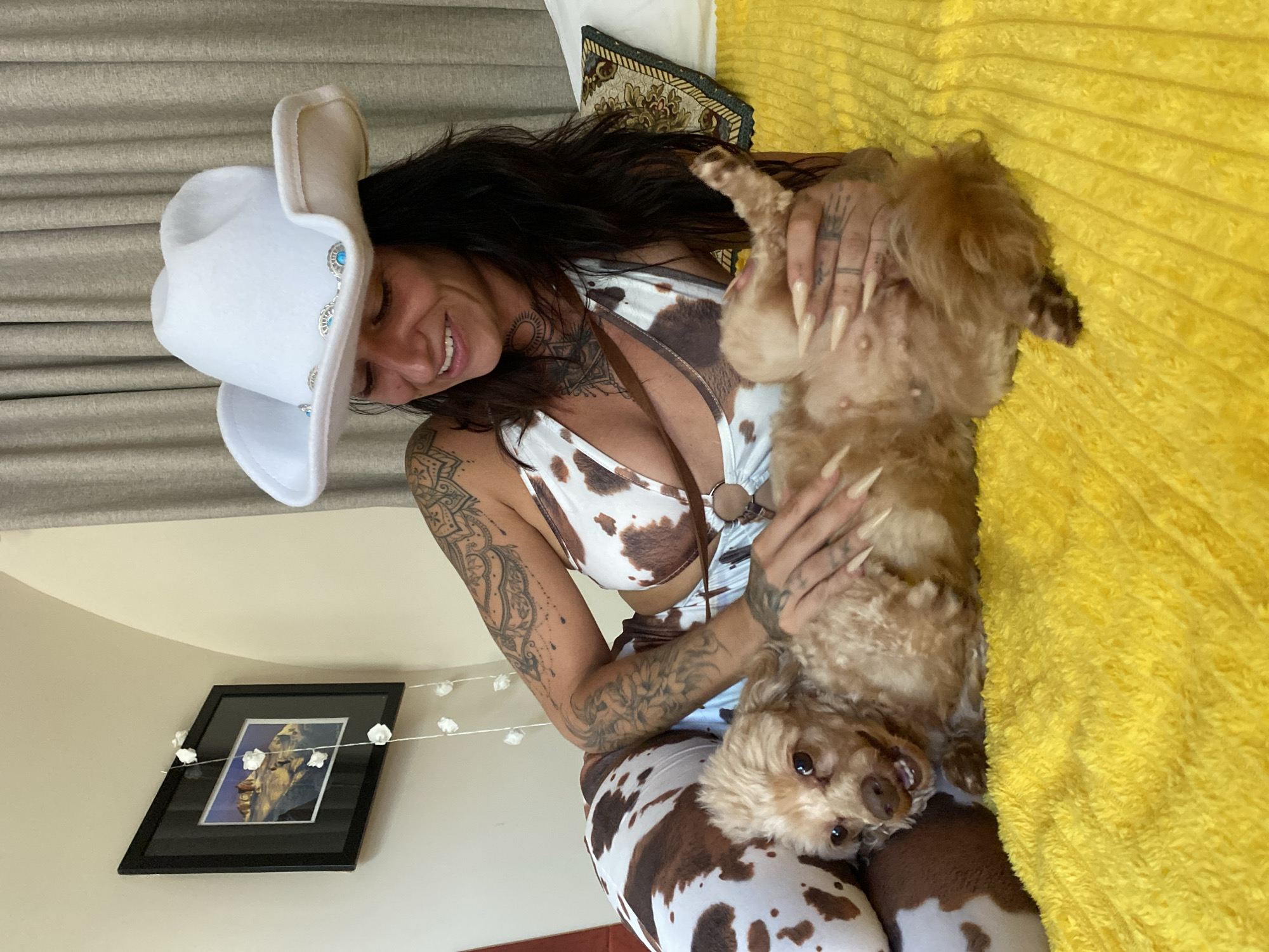 An adult female with long, dark hair and extensive tattoos on her arms and torso is lying on a yellow, textured blanket. She is wearing a white cowboy hat adorned with turquoise stones and a cow-print bikini top. Her left arm is around a small, light brown dog with curly fur, which is lying on its back. The dog's legs are in the air, and it appears to be smiling with its mouth slightly open. The woman's long, light-colored fingernails are visible as she strokes the dog. The background shows a wall with a framed picture and curtains. This scene is not identifiable as being located in Mũi Né, Vietnam.