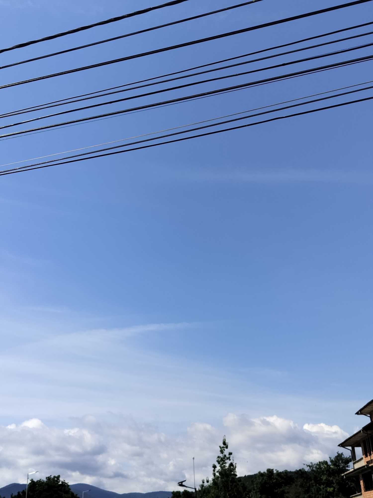 The image captures a bright blue sky with scattered white clouds, seen from a low angle. Several black power lines stretch across the top of the frame. In the distance, a treeline and the outline of mountains are visible. On the right side, a portion of a building with a brown tiled roof and brickwork is present. The setting appears to be the city of Pavel Banya, Bulgaria, on a clear day. There are no people or animals depicted in the scene.