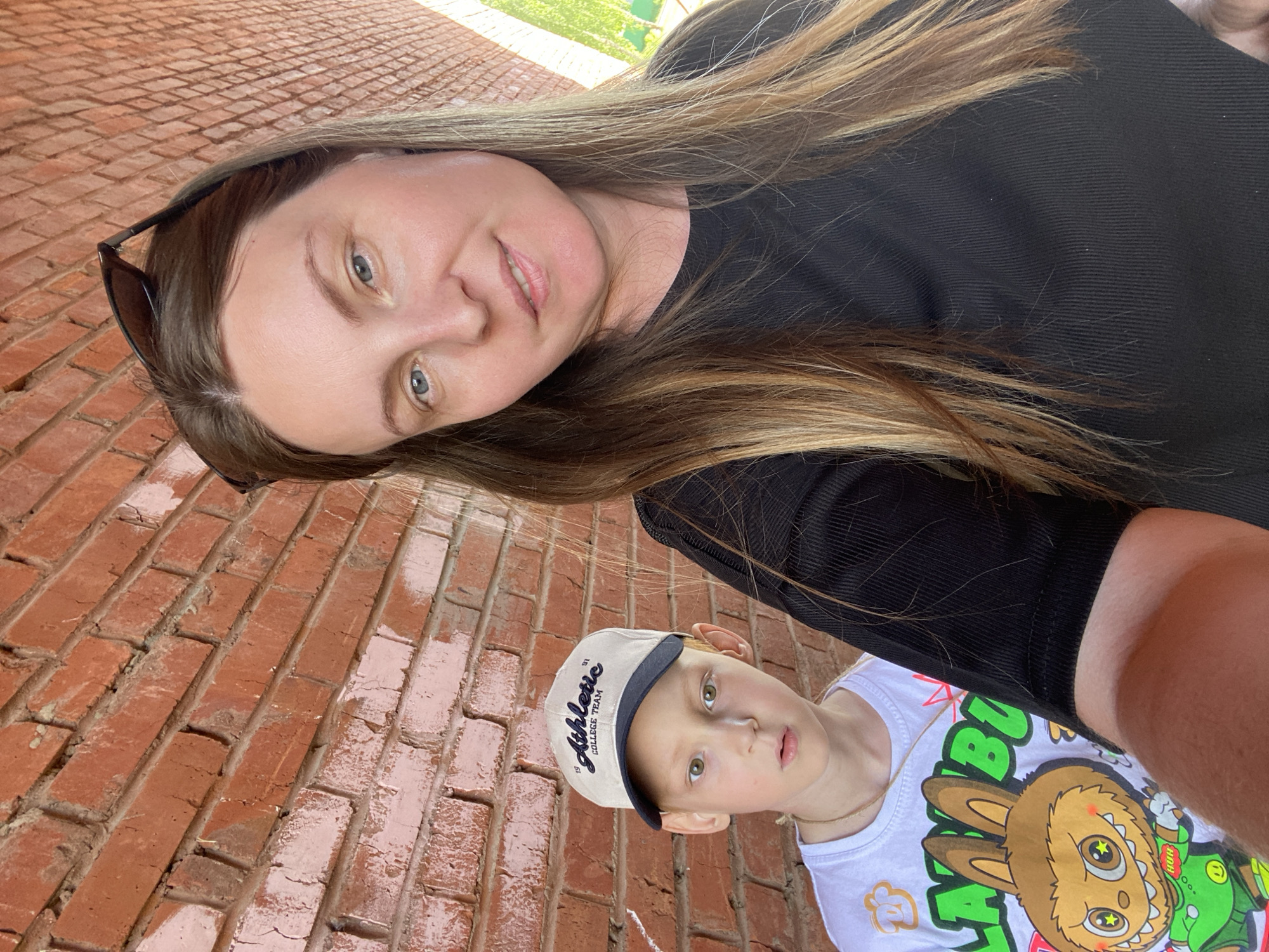 A selfie taken from a low angle shows an adult woman and a child outdoors. The woman has fair skin, light brown hair with blonde highlights, and is wearing a black top. Sunglasses are perched on her head. The child, positioned closer to the bottom of the frame, has fair skin, blonde hair, and is wearing a white baseball cap and a t-shirt with a cartoon graphic. The background appears to be a brick surface. The location could be Noviy, Russia.