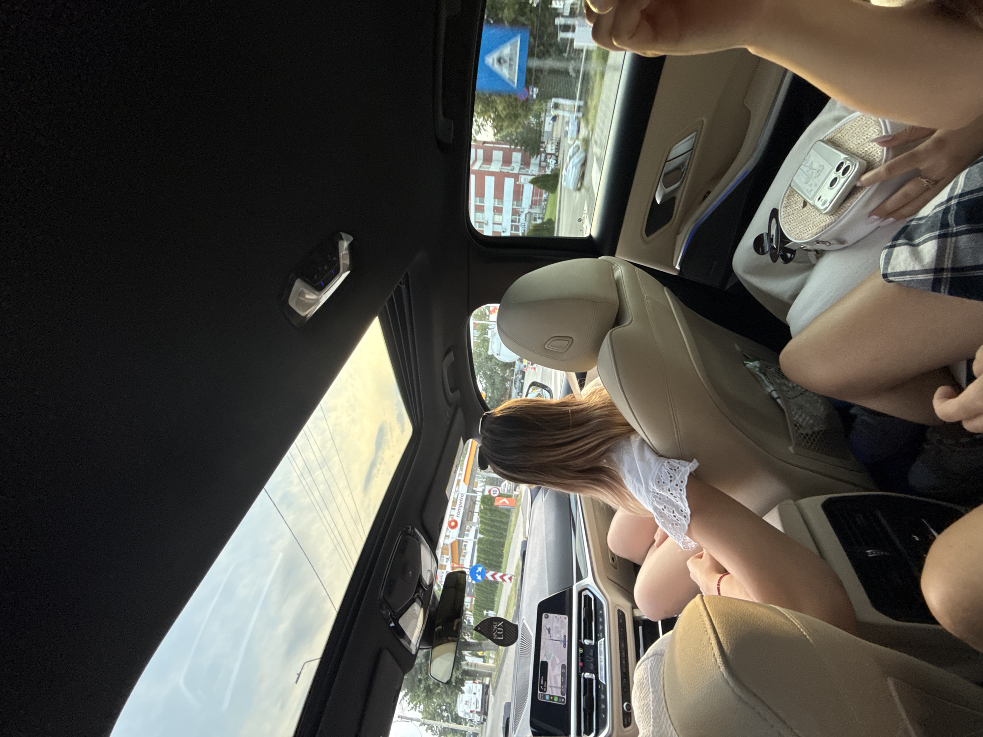 The image captures the interior of a car with a sunroof open, showing a view of a city street in Onești, Romania. Two women are visible, one in the front passenger seat and another in the rear. The woman in the rear, facing away from the camera, has long blonde hair and is wearing a white top with eyelet details. Her legs are crossed, and she appears relaxed. A handbag with a phone on top rests on her lap. In the background, through the car's windows and sunroof, a gas station with "Rompetrol" branding is visible, along with buildings and street signs, indicative of an urban environment. The lighting suggests it is late afternoon or early evening.