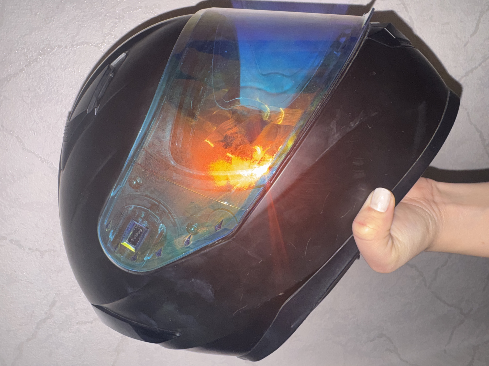 A hand holds a black motorcycle helmet with a reflective, iridescent visor against a textured, gray wall in Bishkek City, Kyrgyzstan. The helmet is positioned at an angle, obscuring most of the viewer's perspective of the wall. The visor displays bright orange and blue hues, reflecting light. The hand, with painted fingernails, is gripping the lower portion of the helmet. The background is a plain, neutral-colored wall with a subtle pattern.