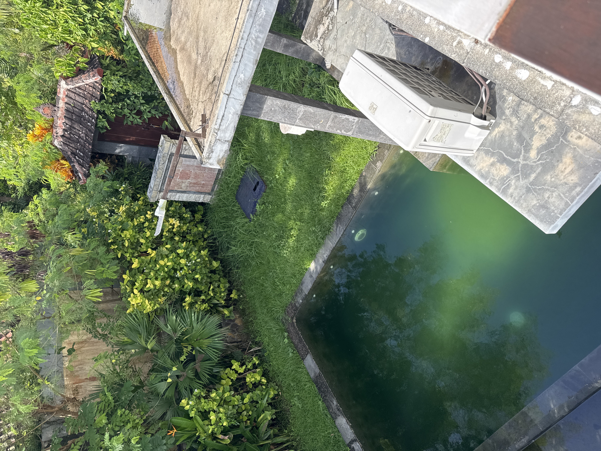 This image shows an elevated view of a building structure and a pool in Kuta Selatan, Indonesia. The pool contains green-tinted water and is bordered by concrete edges and a strip of grass. The building structure has a light-colored roof and concrete supports. An air conditioning unit is mounted on the concrete. Lush vegetation, including trees and bushes, surrounds the building and the pool.