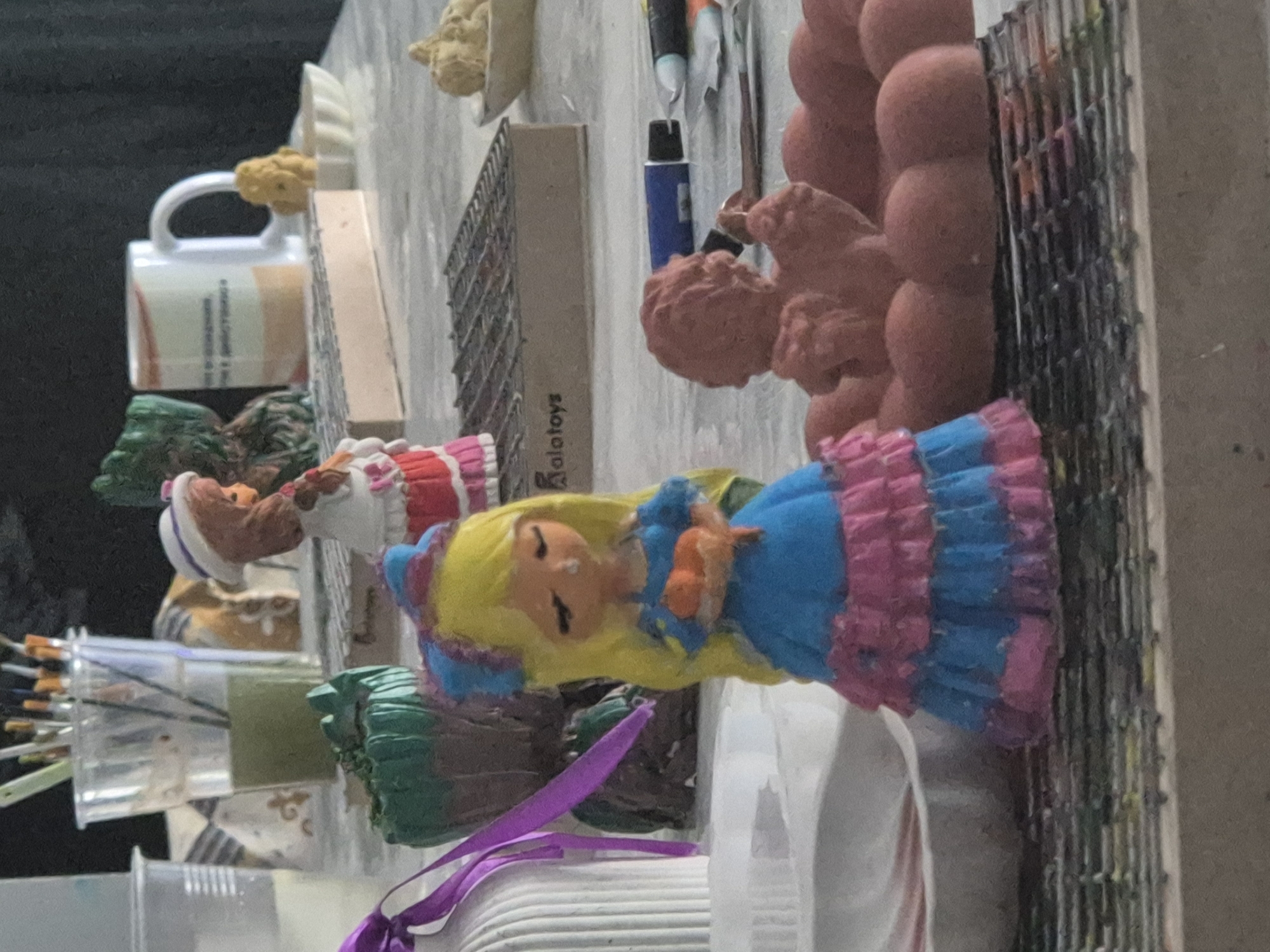 The image depicts a close-up, slightly blurred view of several small, painted figurines and craft supplies arranged on a flat surface. In the foreground, a blonde-haired doll in a blue and pink ruffled dress is holding two orange objects. To the right of the doll is a small, pink, cherubic figure seated on a rounded surface. Behind the doll, there is a green, textured object and a white mug with text on it. Various paint brushes are visible in a clear plastic cup to the left. Several tubes of paint and a blue marker are also present on the surface. The background is dark and out of focus, suggesting an indoor setting. There is no information in the image that allows for the identification of a specific city, including Kokshetau, Kazakhstan.