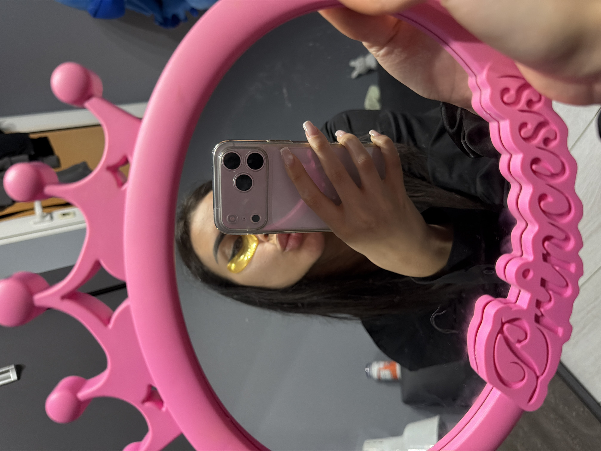 A person is seen taking a mirror selfie in an indoor setting. The reflection shows a female individual, holding a light pink or purple smartphone with a clear case and a triple-lens camera setup, used to capture the image. The person has long, dark hair and is wearing yellow eye patches beneath their eyes, indicating a beauty or self-care routine. Their lips are slightly pursed. They are dressed in a black top.
The mirror itself is a notable object, featuring a distinctive pink frame. The left side of the frame is adorned with a crown-like design, complete with rounded ball embellishments. The right side of the frame displays intricate, raised scrollwork and floral patterns, including a prominent rose motif at the bottom. Cursive text, spelling "Princess," is embossed along this decorative side of the frame.
The background reflected in the mirror appears to be a simple indoor environment with grey walls. A light-colored surface, possibly a table or shelf, is partially visible on the left, and a blue object can be seen in the upper left corner of the reflection. White floor tiles are visible at the bottom of the scene. The image captures a personal moment of self-portraiture within what appears to be a private room in Burgas, Bulgaria.