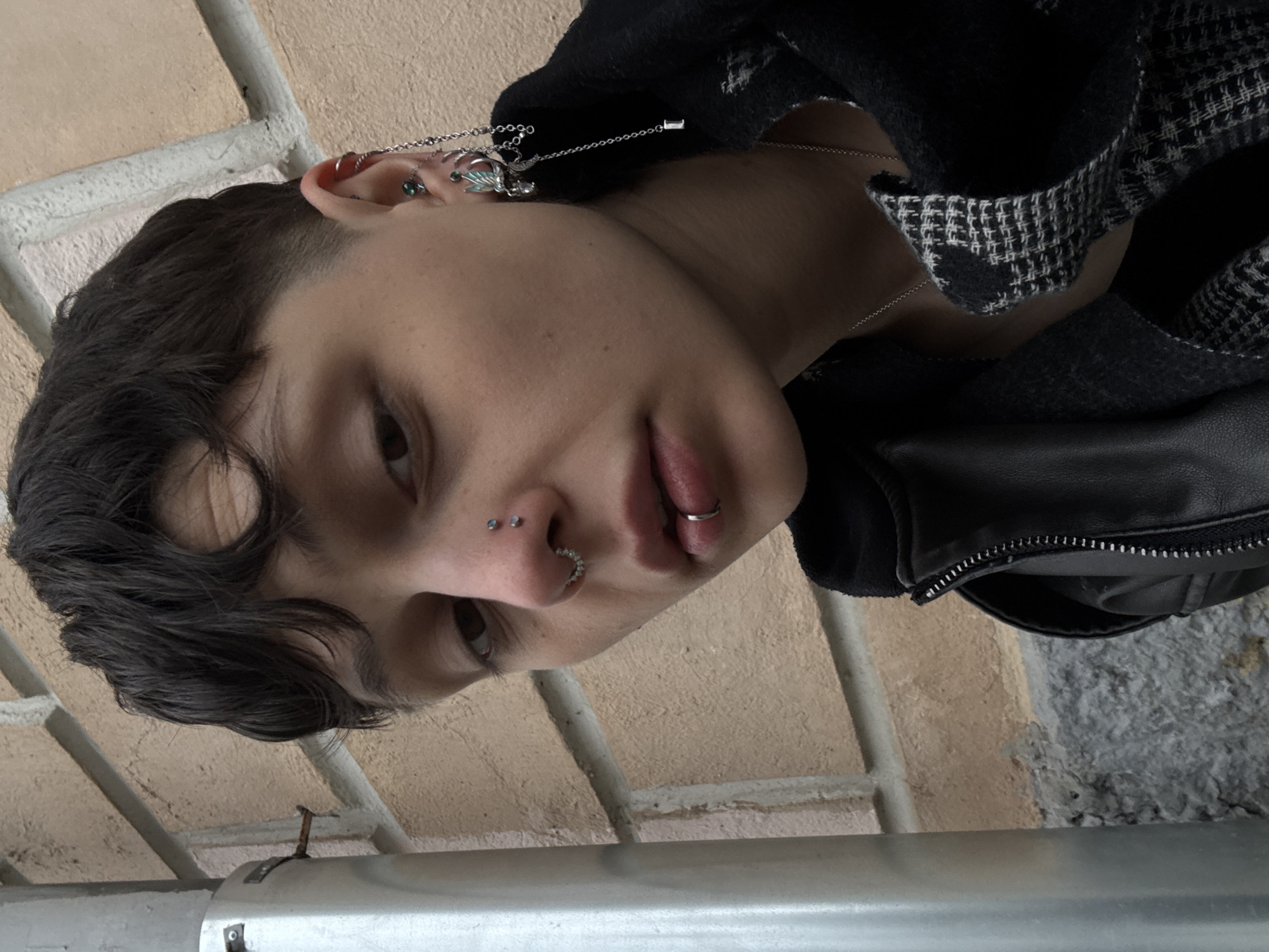 A person with short dark hair and multiple facial piercings, including nose and lip rings, gazes downward. The individual is wearing a dark jacket with a patterned lining. Behind the person is a light-colored brick wall. The location cannot be determined to be Moscow, Russia based solely on this image.