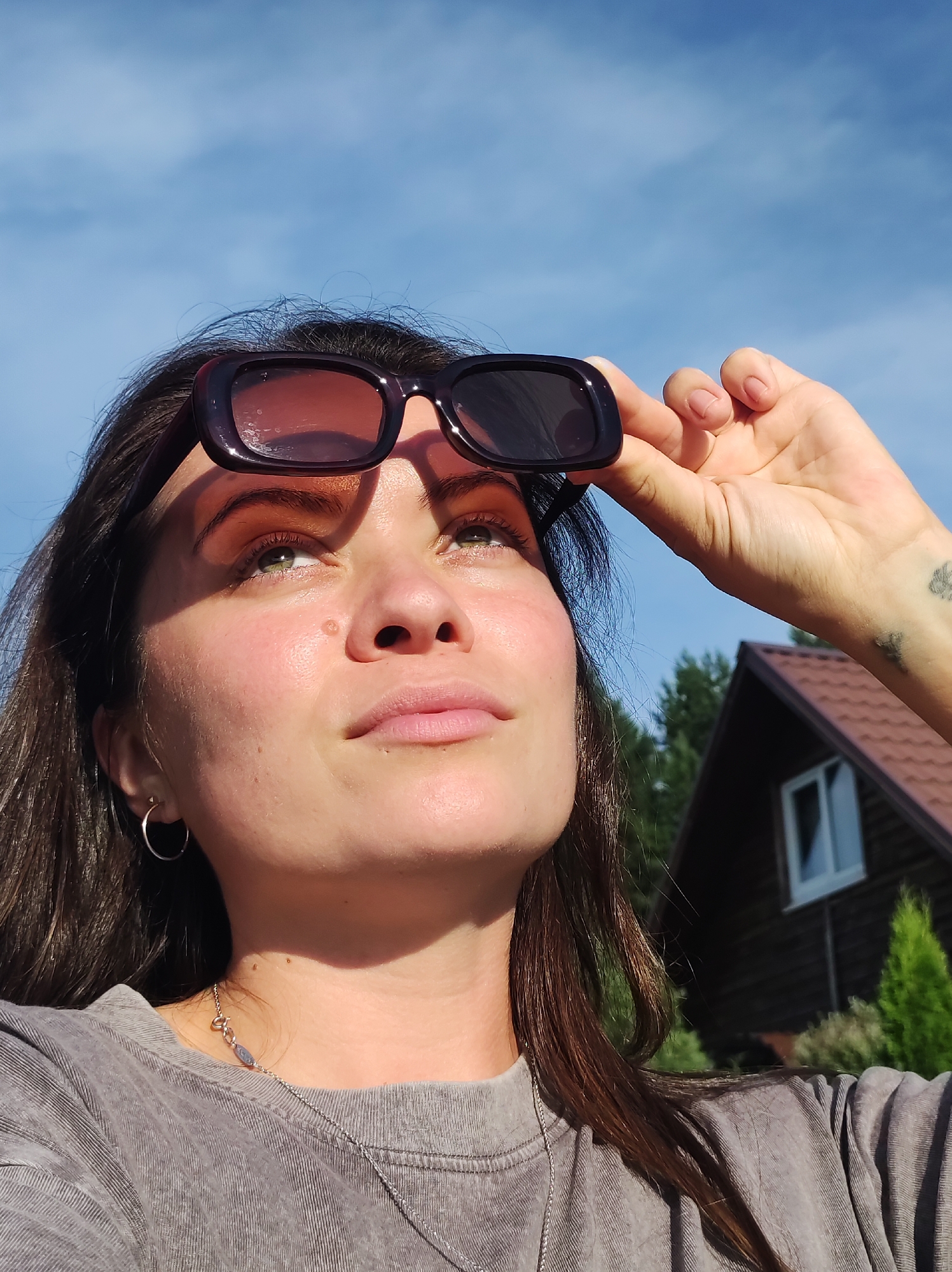 A woman is captured in a close-up, low-angle shot, looking upwards towards a bright blue sky with scattered clouds. She has dark brown hair and is wearing dark-rimmed sunglasses, which she is holding up with her right hand. Her skin is illuminated by the sun, casting a shadow across her face. She is wearing a grey t-shirt and a silver necklace. In the background, a wooden house with a red roof and a white-paned window is visible, surrounded by greenery and trees. The setting appears to be a rural or suburban area, possibly on the outskirts of the city of Fokinskoye, Russia.