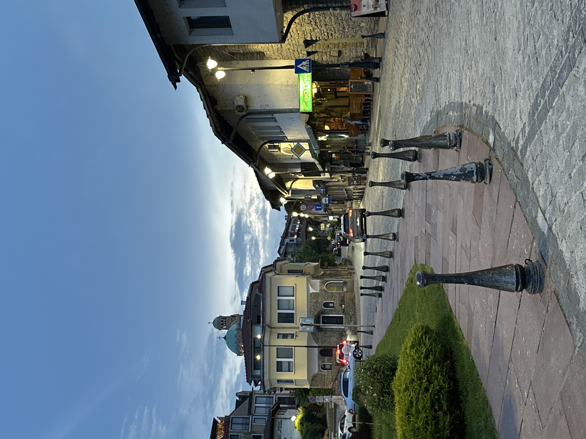 This media file presents a street scene in Veliko Tarnovo, Bulgaria, captured during dusk or early evening. The image, rotated 90 degrees clockwise, displays a vibrant street lined with traditional European-style buildings under a clear blue sky dotted with scattered clouds.
On the left, a row of buildings with stone and painted facades features multiple windows and balconies. Streetlights are illuminated, casting a warm glow that highlights the architectural details and the paved street. A prominent green neon sign for "GOTVIN Market" stands out above a shop entrance. Other visible signage includes a blue pedestrian crossing sign, a yellow diamond sign (often indicating a priority road), and smaller blue road signs, such as a circular one-way arrow. A partial "COSTA" sign is also discernible.
The street and pedestrian areas are paved with a mix of cobblestones and reddish pavers. A series of distinctive dark, decorative bollard-like structures, resembling stylized cannons, line the edge of a grassy median adorned with manicured bushes. In the background, additional buildings are visible, most notably one with a striking green/teal dome topped with a cross, likely identifying it as a church. Cars can be seen on the street, some with headlights on, suggesting ongoing traffic. No people are explicitly visible in the frame, contributing to a calm and atmospheric evening scene.