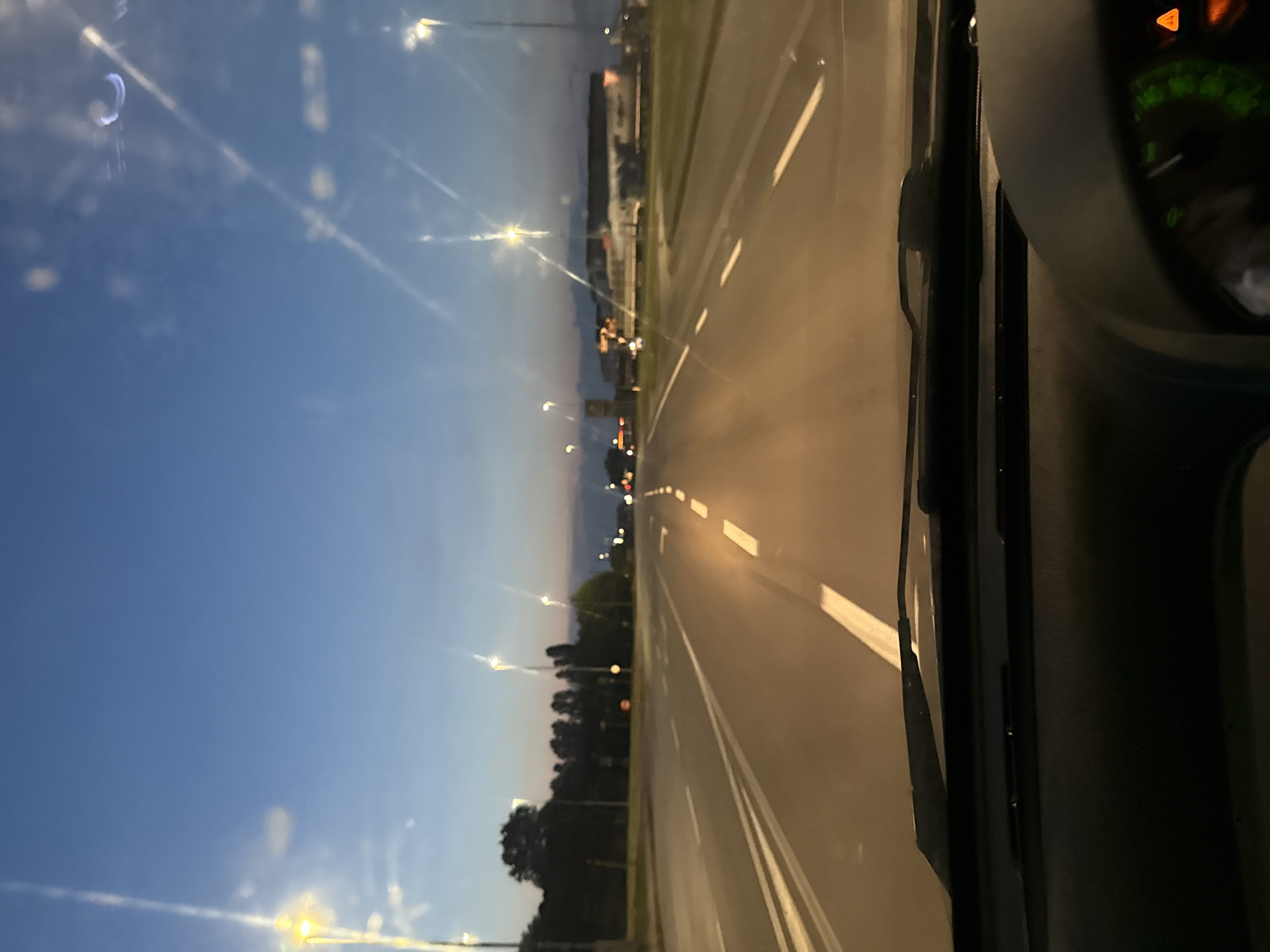 The image captures a view from inside a vehicle driving on a road at dusk, possibly in Casalmaggiore, Italy. The road features clear lane markings, with streetlights illuminating the path. Trees line the side of the road, and in the distance, there appears to be a commercial or industrial area with buildings and vehicles. The sky is a deep blue, transitioning from lighter hues near the horizon. The dashboard of the vehicle is partially visible in the foreground.