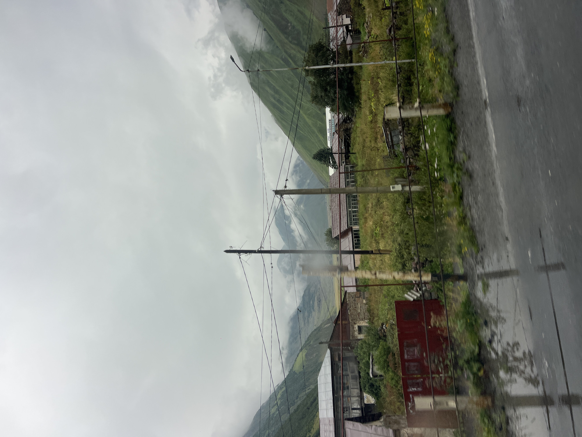 The image depicts a cloudy, overcast day in a rural mountainous area, likely Almasiani, Georgia, given the context. The foreground features a wet asphalt road, reflecting the sky and the surrounding environment. A metal fence runs horizontally across the image, separating the road from a grassy area dotted with small yellow flowers.
Behind the fence, several structures are visible. To the left, a long, low building with a corrugated metal roof stands. Adjacent to it is a smaller, older-looking building constructed with stone and wood, featuring a partially collapsed section. Further to the right, another building with a similar metal roof is present, along with a small, weathered shed. The vegetation in this area is lush and green, with trees and bushes growing around the buildings.
In the mid-ground, power lines crisscross the scene, supported by utility poles. One pole, made of wood, stands prominently in the right half of the image. Another utility pole, also wooden, is visible on the left. In the background, majestic green mountains rise, their peaks shrouded in mist and clouds, creating a dramatic and atmospheric landscape. The overall impression is one of a remote, somewhat rugged, and atmospheric setting. There are no people visible in the image.