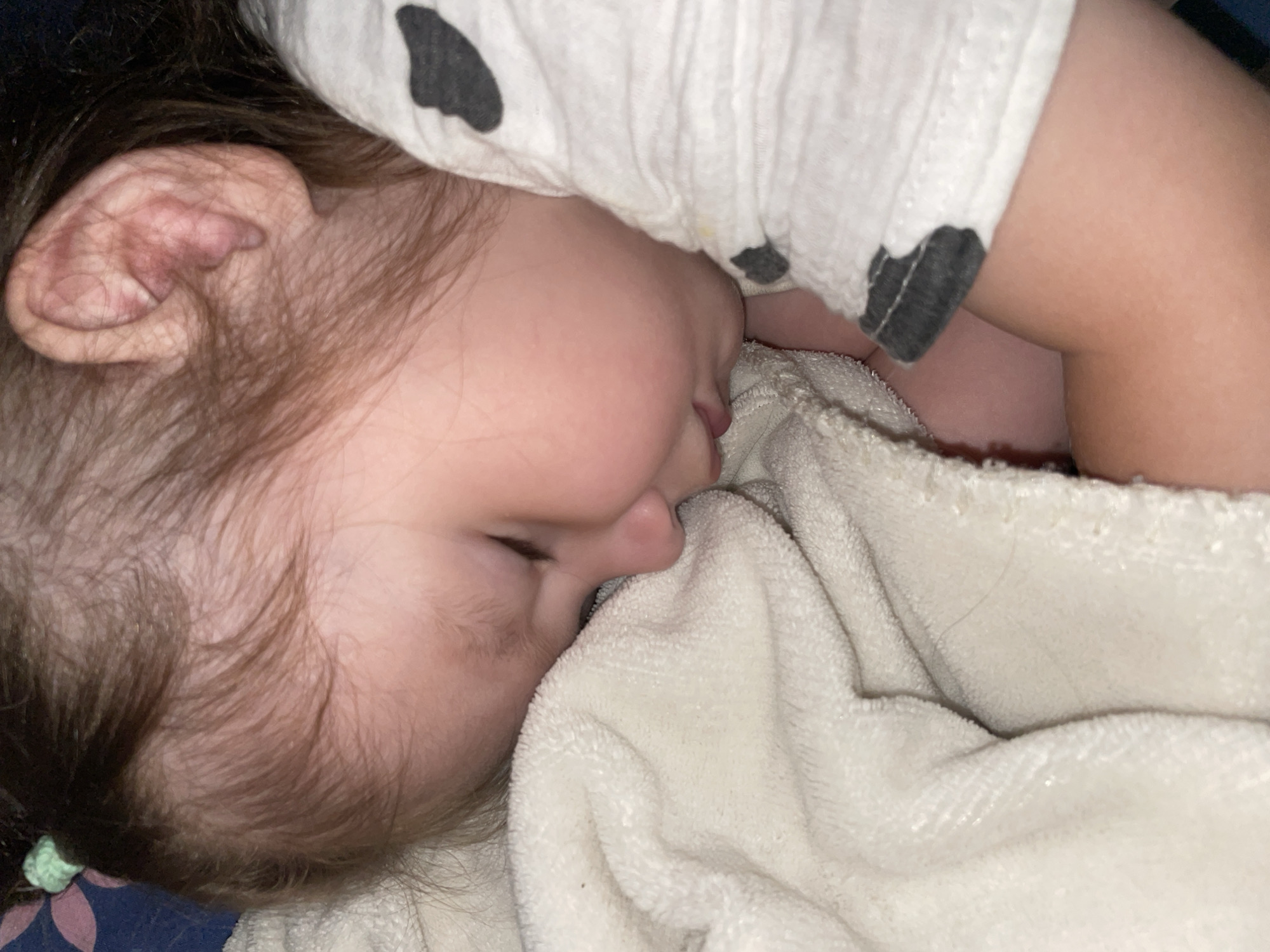 A close-up image depicts a young child, likely an infant or toddler, sleeping peacefully indoors. The child's face is shown in profile, turned towards the right, with their eyes closed and a serene expression. Their left ear is clearly visible, with fine, light brown hair framing their face. A small green elastic or tie can be seen in their hair near the lower left of the frame.
The child is partially nestled into a soft, light-colored (possibly white or cream) towel or blanket, which covers their mouth and nose area, contributing to a sense of comfort and warmth. They are wearing a white garment, possibly a top or a onesie, adorned with several dark, irregularly shaped spots, giving it a playful, perhaps cow-print, design. A portion of the child's arm is visible on the right, also resting on the blanket.
The lighting in the image is dim, suggesting a nighttime setting or a subdued room environment, ideal for sleep. The atmosphere is quiet and intimate, focusing solely on the sleeping child. The image provides no specific architectural or environmental cues to indicate the provided location context of Izhevsk, Russia. There is no visible text in the image.