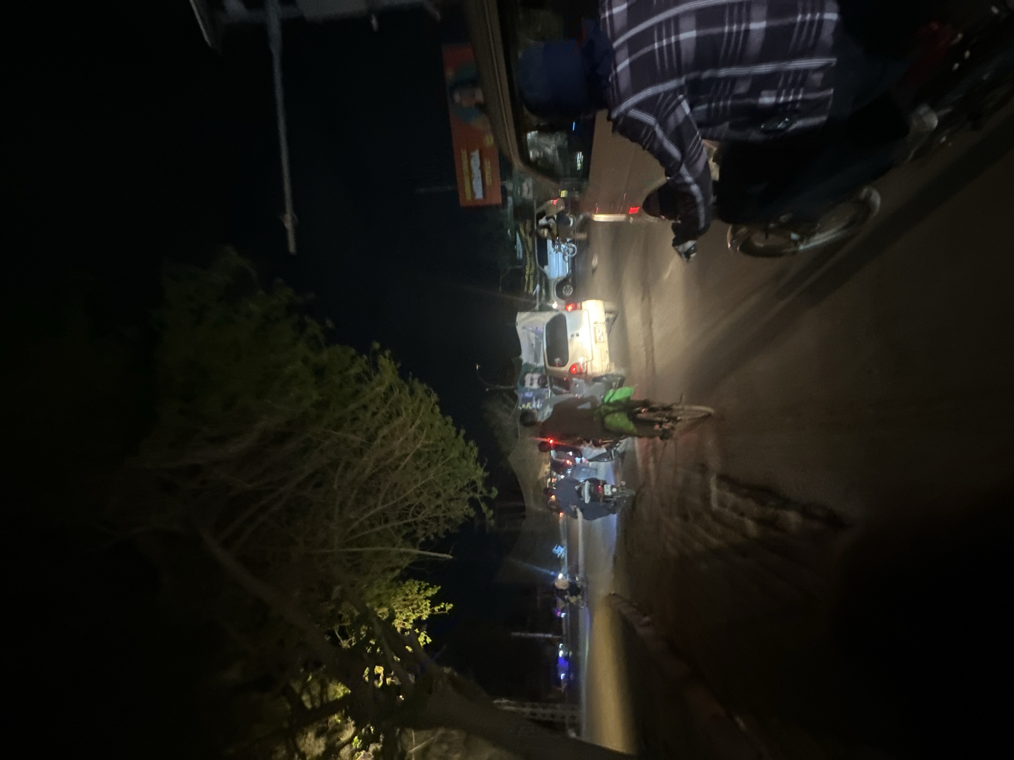 The image shows a street scene at night in Quetta City Tehsil, Pakistan. Traffic is moving along a paved road, illuminated by vehicle headlights and streetlights in the distance. A person wearing a dark jacket and a plaid shirt is riding a scooter or motorcycle on the right side of the road. On the left of the road, a tree with foliage is visible. A billboard is partially visible in the background. Several other vehicles, including cars and motorcycles, are present on the road. The overall lighting suggests it is nighttime.