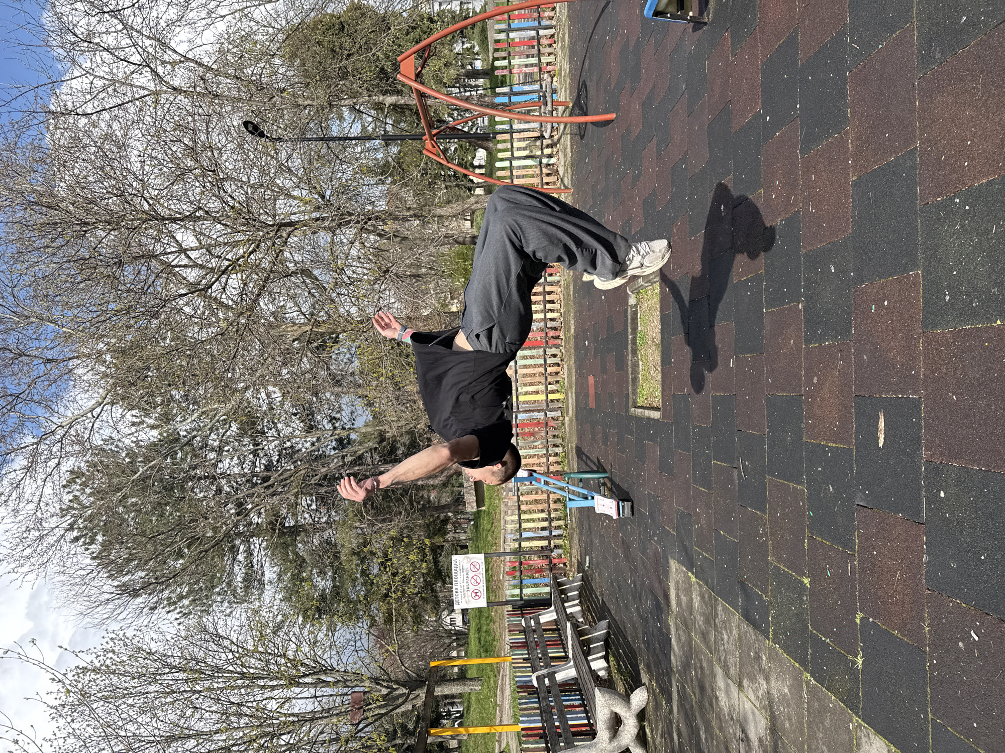 The image shows a man performing a backflip in a playground setting. The playground is located in Shumen, Bulgaria, as indicated by the sign written in Bulgarian. The man is wearing a black t-shirt, gray pants, and white sneakers. He is in mid-air, with his body arched backward. The playground has a safety surface made of interlocking rubber tiles in brown and black. In the background, there are trees with bare branches, and playground equipment including an orange A-frame climbing structure and a colorful fence. A bench is visible in the lower right corner. A sign with warnings is mounted on the fence. The lighting suggests it is daytime.