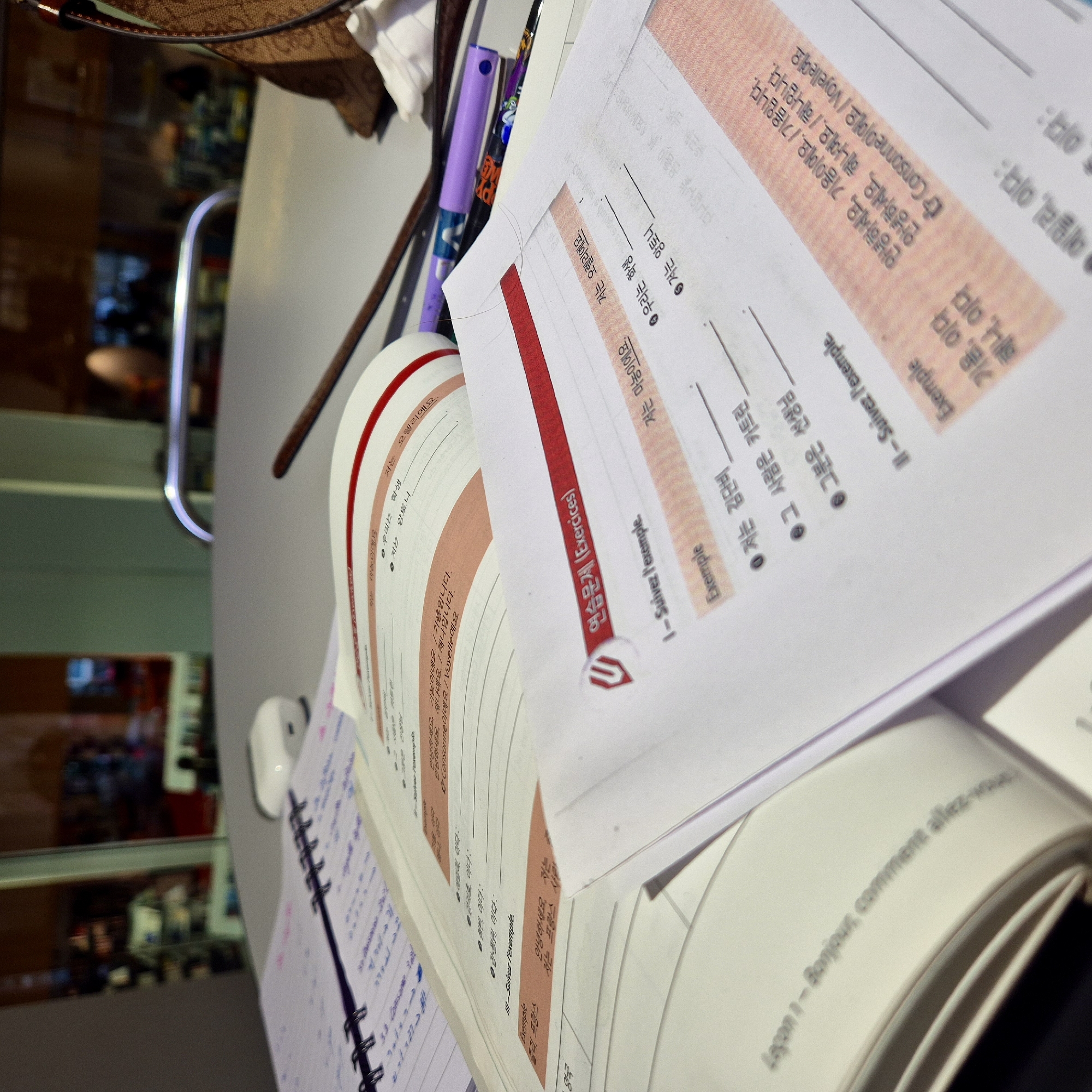 The image shows an open notebook and textbook, seemingly on a desk. The notebook has spiral binding and handwritten notes. The textbook appears to be Korean and French language learning material, opened to exercises and examples. Pens are scattered around the book. The backdrop is a blurred reflection indicating a modern interior setting.