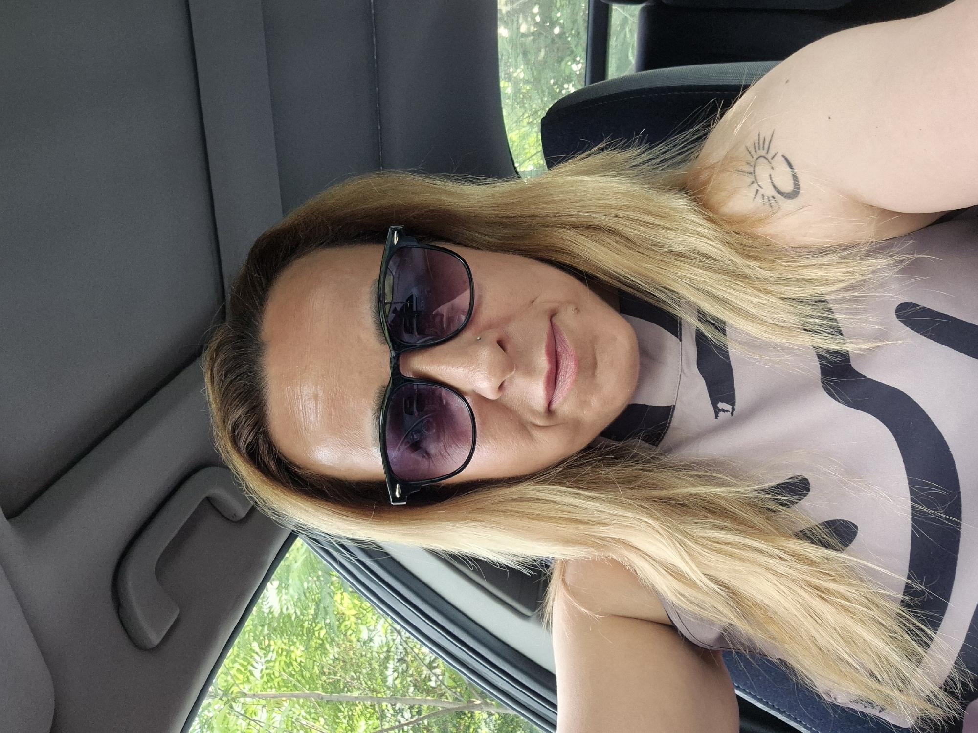 A female individual, appearing to be an adult, is depicted in a close-up, chest-up shot, seated inside a vehicle. Her light brown hair, with blonde highlights, extends past her shoulders. She is wearing dark-framed sunglasses with purple-tinted lenses. A small silver stud is present in her left nostril. Her lips are a light pink color. A small, dark mole is visible on her upper lip, to the left of her philtrum. On her left upper arm, a black tattoo depicting a stylized sun and crescent moon is visible. She is wearing a sleeveless top with a light beige or grey background and a repeating dark, abstract pattern. The car's interior features a grey headliner and a grey grab handle above the passenger-side window. Green foliage, indicative of trees and bushes, is visible through the side and rear windows of the vehicle, suggesting an outdoor daytime setting. The individual is positioned to face the camera directly, suggesting a self-portrait. The location is an unknown city.