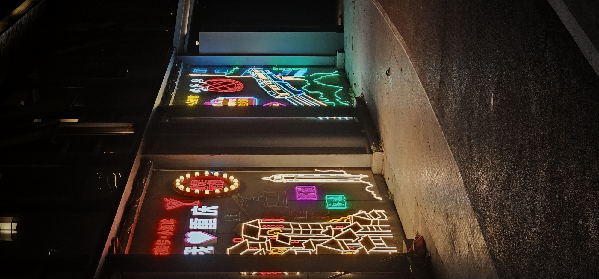 This nighttime image captures two vibrant neon sign installations, vertically stacked, illuminating a dark, urban environment, likely a passage or building facade. The perspective is tilted, looking upwards at the glowing displays against the deep shadows of surrounding structures, including a textured concrete wall on the right.
The upper neon sign features a dynamic array of colors and shapes. It includes a prominent green outline resembling a geographical map or city landscape, intertwined with blue and white linear elements that could represent roads or waterways. A red, grid-patterned oval object and partially visible yellow Chinese characters add to the composition, suggesting local cultural or commercial themes.
The lower and more elaborate sign is a tapestry of glowing light. On the left, an oval-shaped display with numerous small white bulbs encircles red Chinese characters, which could signify "日月" (sun, moon) or a similar phrase. Below this, bright red and white neon characters are visible, clearly spelling out "江北小吃" (Jiangbei Snacks or Eatery), accompanied by a white heart shape. To the right, a large, intricately outlined traditional Chinese architectural structure, perhaps a temple or historical building, glows in yellow, white, and red. Nearby, smaller pink and green square neon signs contain white characters. A long white neon outline resembling a stylized brush or column is also part of this detailed scene.
The image portrays a lively and modern urban atmosphere, highlighting the unique visual culture of nighttime cityscapes. The choice of traditional architectural elements and the explicitly readable Chinese text "江北小吃" strongly indicate the setting as 江北城街道, China, showcasing local identity through contemporary art and advertising.