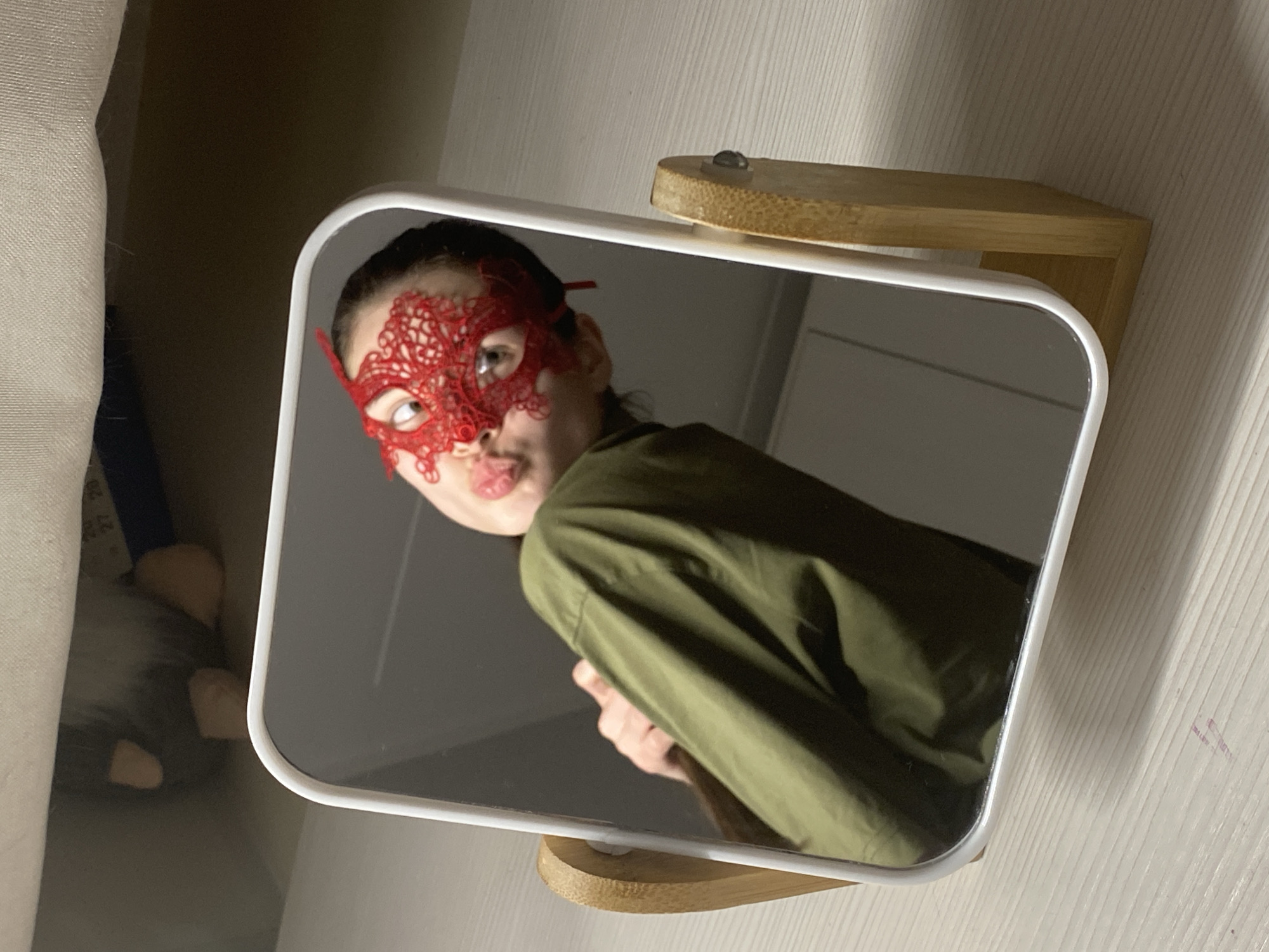 A person's reflection is captured in a rectangular mirror with rounded corners, set on a light-colored wooden stand. The mirror and its reflection are tilted at an angle, presenting a skewed view.
The reflected person is wearing a prominent red lace mask that covers their eyes and extends over the nose and forehead. Their hair appears to be pulled back, and they are dressed in a dark olive green, long-sleeved shirt or jacket. One eye is clearly visible through the mask, looking towards the camera, while their tongue is slightly out, conveying a playful or cheeky expression. Part of their left hand is visible in the lower part of the reflection.
The background visible through the reflection is dark and indistinct, suggesting an indoor setting. Outside the mirror's frame, on the left, a light-colored, textured surface (possibly a wall or fabric) is visible, along with a partially obscured, dark object that appears to be a grey and white stuffed gnome-like figure. Faintly, on a blue object behind the mirror, some numbers like "27" and "20" can be glimpsed. On the right, a light-colored wooden surface is visible, matching the aesthetic of the mirror's stand. The overall lighting suggests an indoor setting, possibly with artificial light.