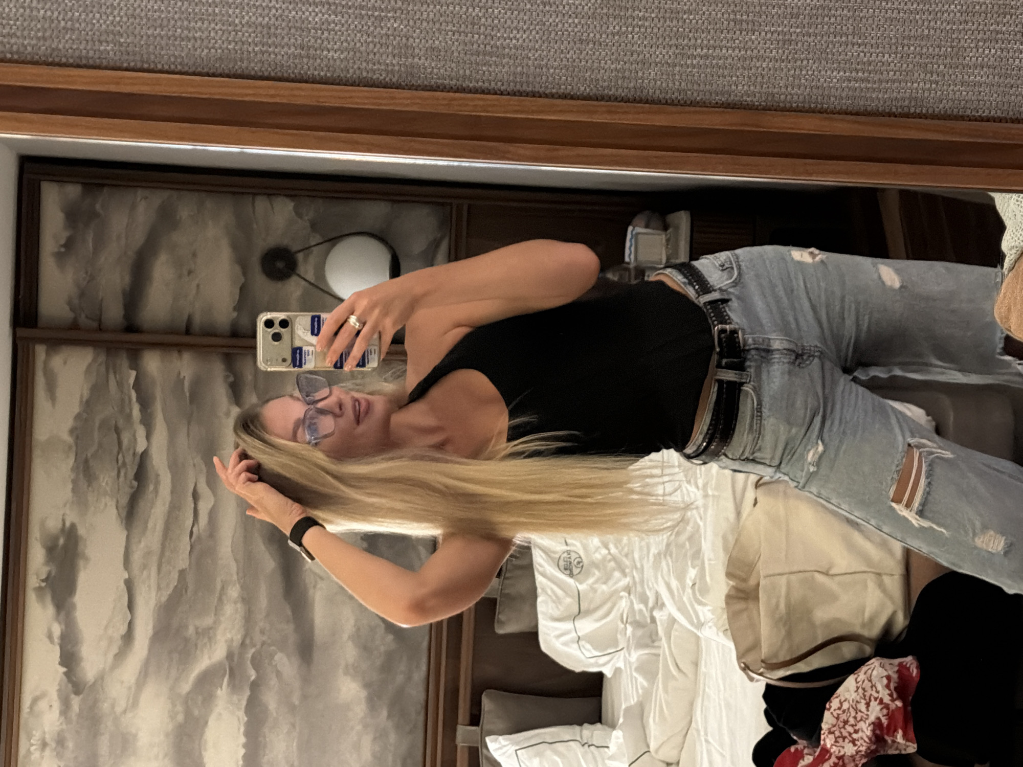 A woman is posing in front of a mirror, taking a selfie with her phone. She has long blonde hair, is wearing glasses, a black tank top, distressed jeans, and a watch. The background reflects a room with a bed, pillows, a decorative wall panel, and the mirror's frame. There is no information in the image to confirm the location in the city of Beldibi, Turkey.