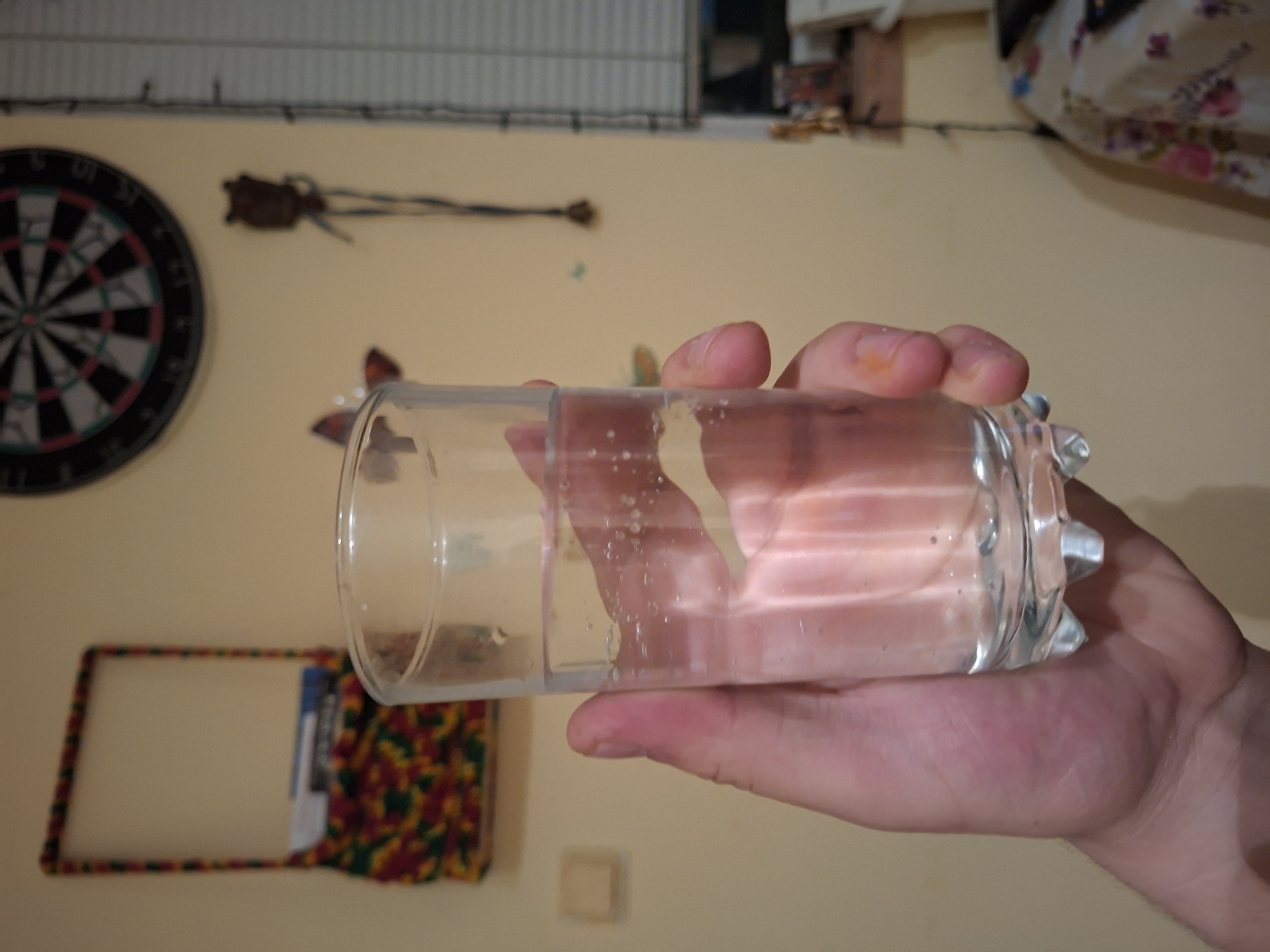 A hand holds a glass of liquid. In the background, a dartboard, a wall decoration, and a framed display are visible on a light-colored wall. The location is not specified.