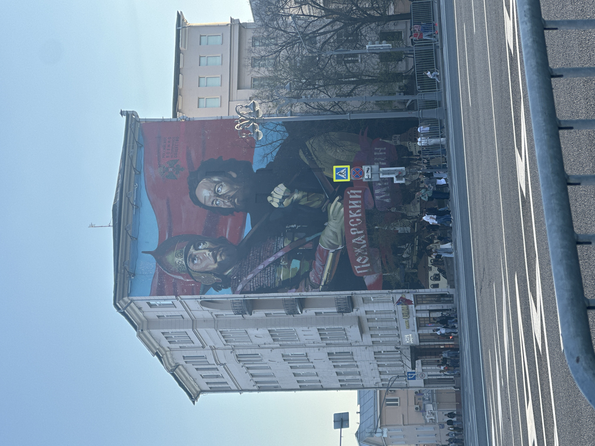 The image shows a large mural on the side of a building in Moscow, Russia, depicting the historical figures Minin and Pozharsky. The mural is painted in a realistic style, featuring the two men prominently against a red and blue background with other figures in the background. The building is multi-storied with light-colored walls and symmetrically arranged windows. In front of the building, a street runs parallel, with white lane markings visible. There are people gathered on the sidewalk near the building, watching something out of view, possibly a parade or event. A grey metal barrier is partially visible in the right foreground of the image, and trees are also visible in the background behind the building. A street sign is seen which is also on the mural. The overall environment is urban with clear sky above.