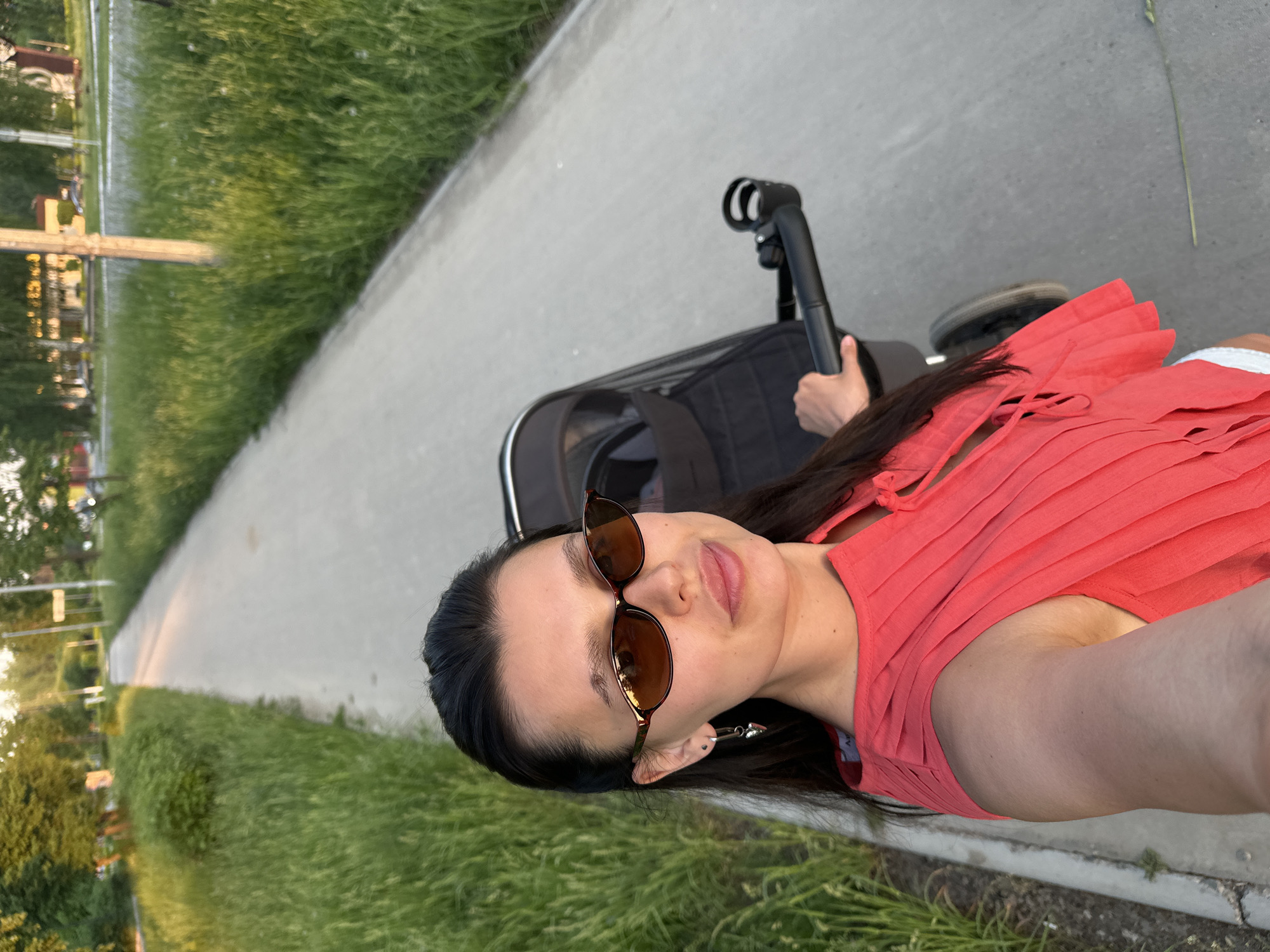 The image shows a woman taking a selfie while walking on a paved path, possibly in a park. She is wearing sunglasses and a red top, pushing a baby stroller with her right hand. The path is bordered by green grass on both sides. There are trees and buildings in the background, which appear to be a part of an urban area. Based on the prompt, this could be the city of Chaikovsky, Russia.