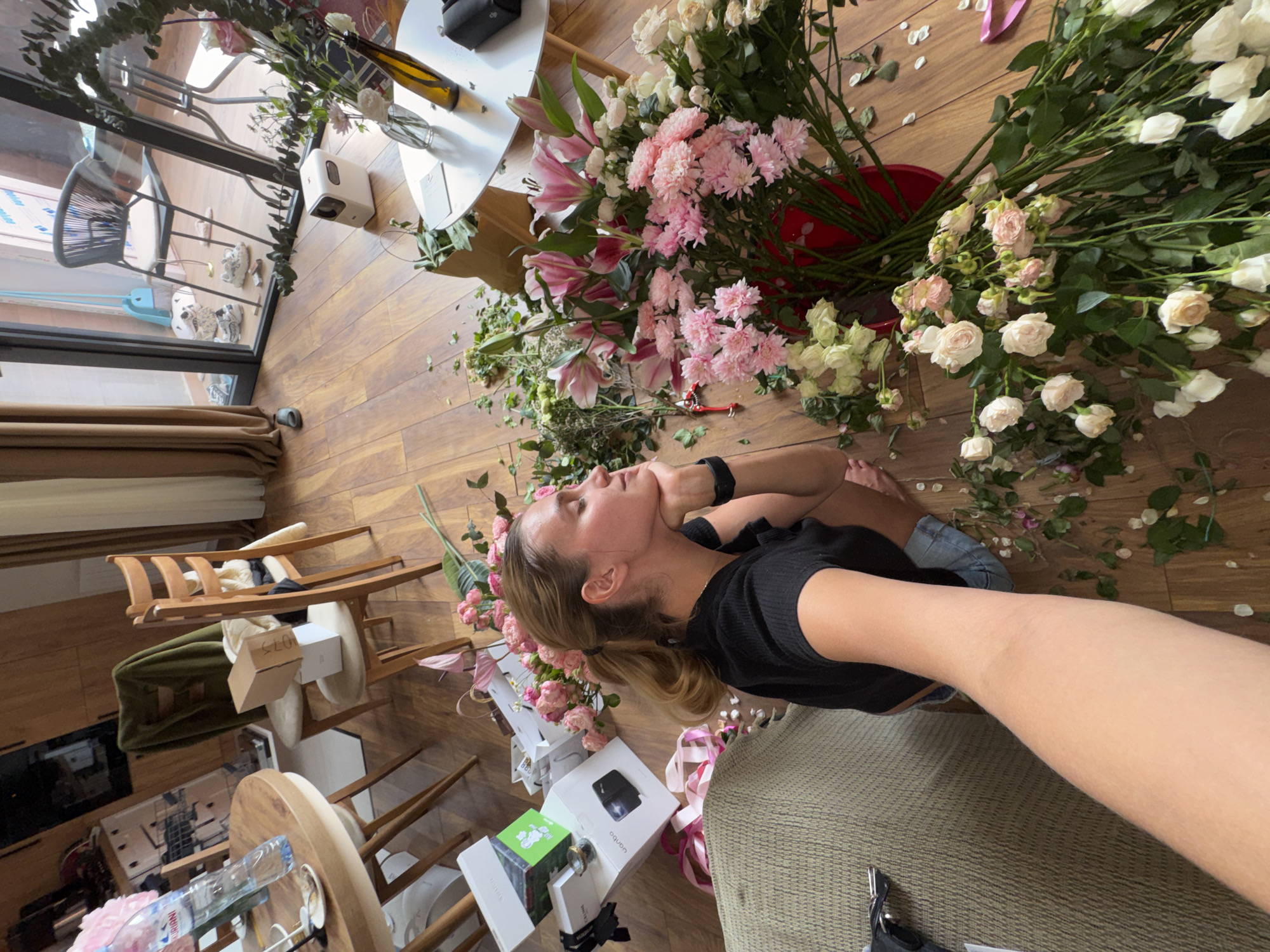 The image, rotated 90 degrees clockwise, displays a young woman surrounded by a large assortment of fresh cut flowers in an indoor setting. The woman, with light brown hair and wearing a black short-sleeved top and light-colored shorts, is lying on her side on a wooden floor. She looks upwards with a calm, pensive expression, her right hand resting near her chin, and a smartwatch visible on her left wrist.
A profusion of flowers covers much of the scene, predominantly pink lilies, pink chrysanthemums, and numerous white and light pink spray roses. Many of the long-stemmed flowers are gathered in a red bucket, while others, along with scattered petals and green leaves, are spread across the polished wooden floor. A pair of red pruning shears lies among the flower stems, suggesting an ongoing process of flower arranging or preparation.
The setting appears to be a bright, modern living space with warm wooden flooring. Natural light streams in through large glass windows or doors visible in the background, which also show glimpses of an outdoor area with a chair and a light blue object. Interior furnishings include a minimalist wooden chair, a round white table holding a small white electronic device (possibly a humidifier) and a dark bottle, and another wooden table with a "FIJI Water" bottle. Several gift boxes, some open, are scattered near the woman, including one with "OQQU" printed on it, alongside pink ribbons. A textured, light-colored fabric surface, likely part of a sofa or cushion, is also present where the woman's leg rests. The scene conveys a tranquil moment amidst a creative floral endeavor.
