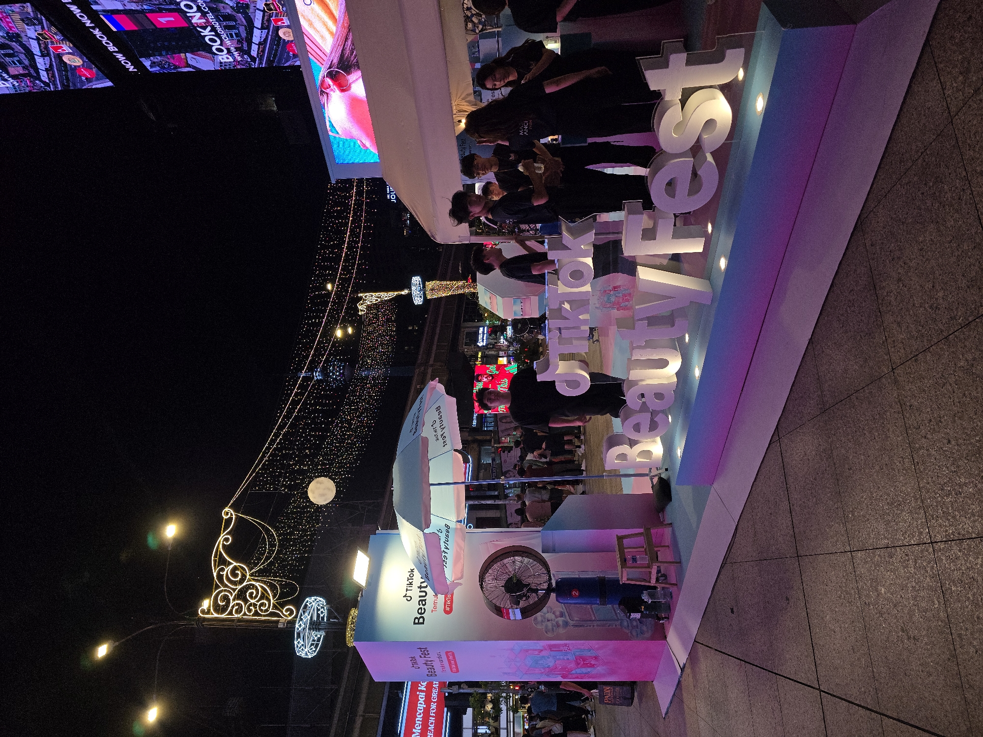The image depicts a nighttime street scene in Kuala Lumpur, Malaysia, likely within a commercial district given the presence of large digital billboards and streetlights. The focal point is a temporary outdoor booth set up for a "TikTok Beauty Fest." The booth features a large, illuminated sign spelling out "BeautyFest" in white 3D letters, with "TikTok" also prominently displayed. The structure is adorned with pink and blue lighting, creating a vibrant atmosphere.
Several individuals, appearing to be staff or participants, are gathered around the booth. They are dressed in dark clothing, with some wearing T-shirts with logos. One person is standing near a large fan, another is holding a bottle, and a group is conversing near the tented area of the booth. The background reveals more of the urban environment, including streetlights, decorative string lights, and the bright displays of digital billboards. A large, illuminated full moon is visible in the night sky. The ground appears to be paved with large, square tiles. The overall scene suggests an event in progress, with an emphasis on the "TikTok Beauty Fest" branding and the commercial backdrop of the city at night.